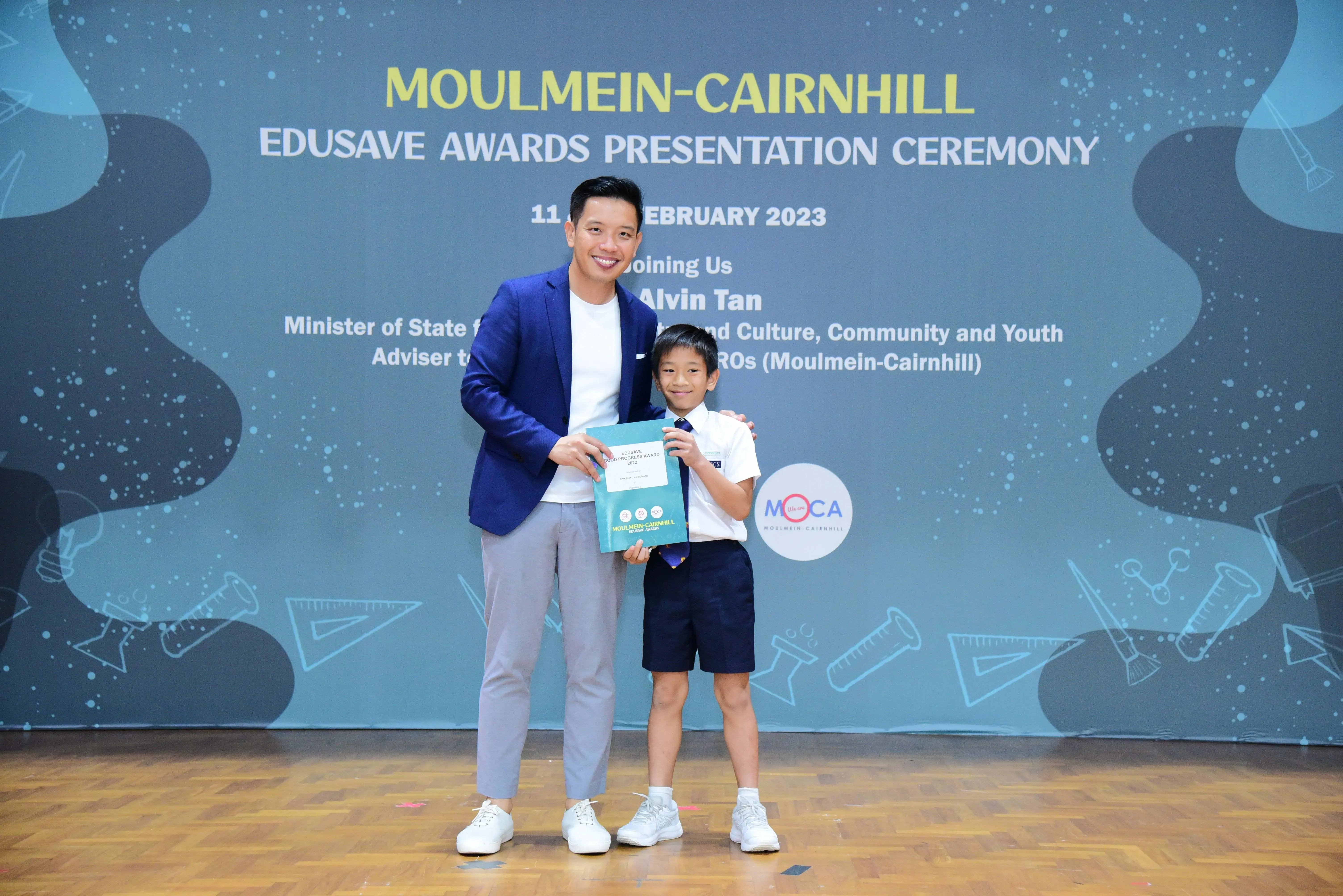 A man in a blue blazer and a young boy in school uniform pose with an award at the Moulmein-Cairnhill Edusave ceremony.