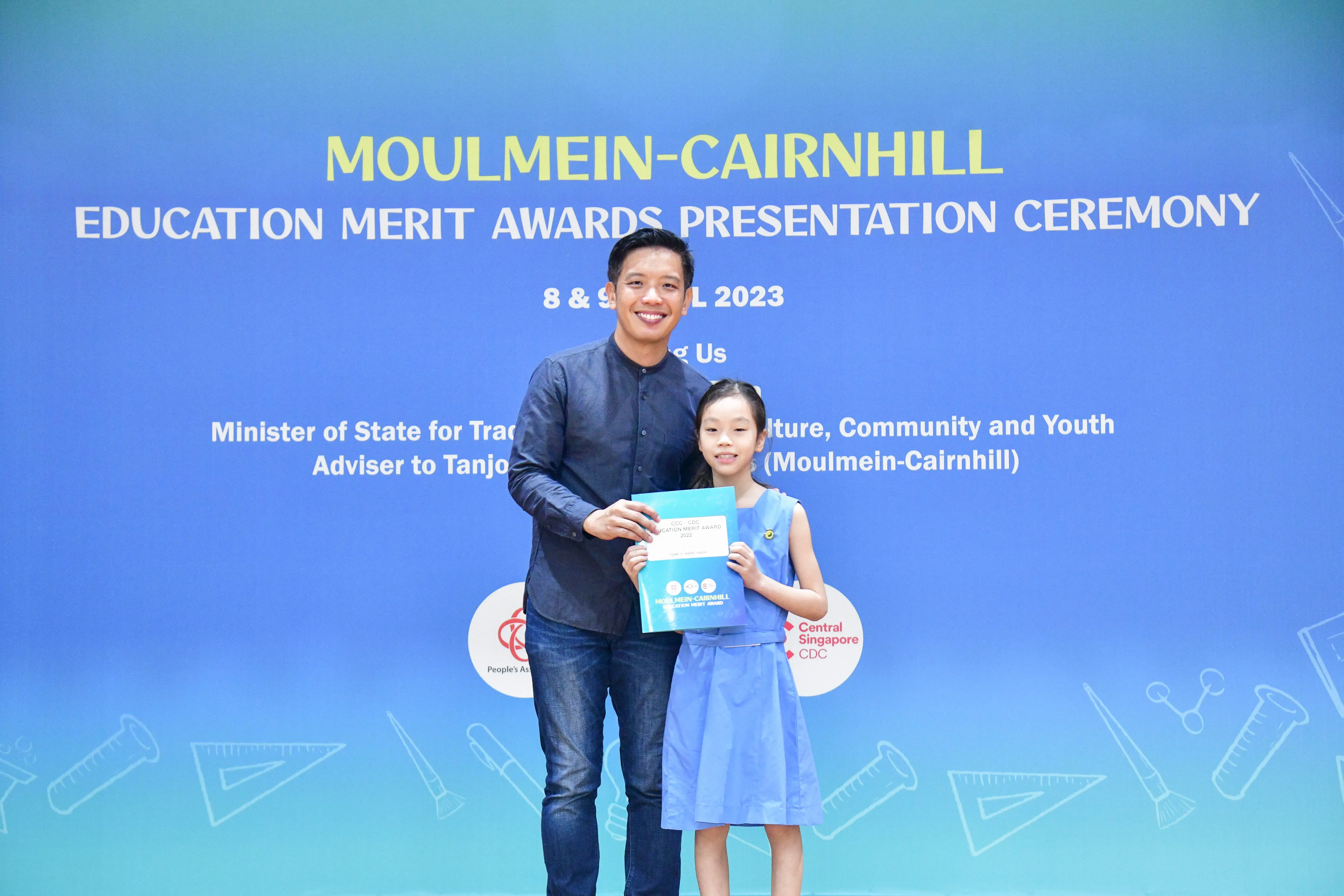 A man and a young girl hold a certificate together in front of a Moulmein-Cairnhill Education Merit Awards 2023 backdrop.