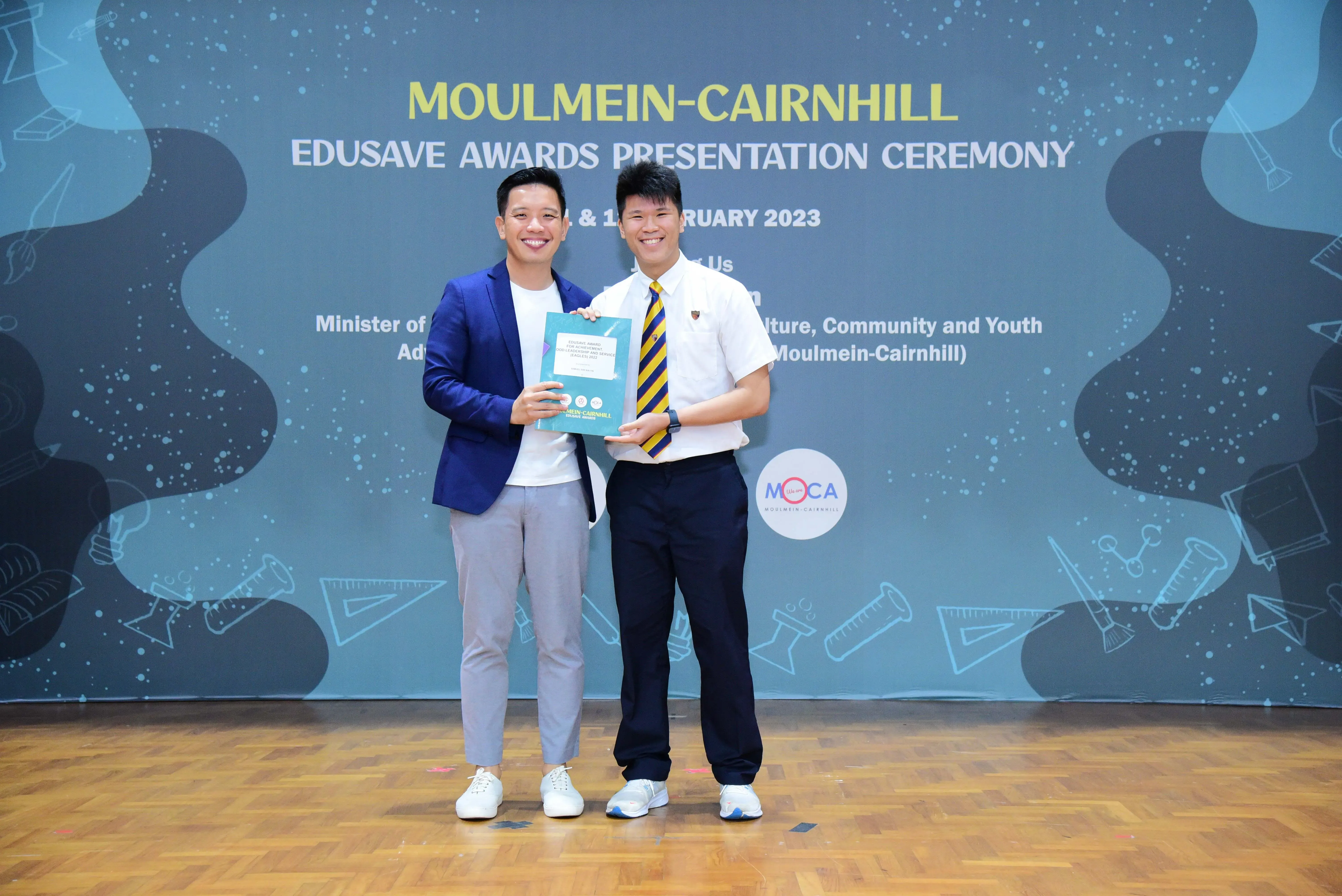 Two smiling men hold a certificate at the Moulmein-Cairnhill Edusave Awards Presentation Ceremony 2023.