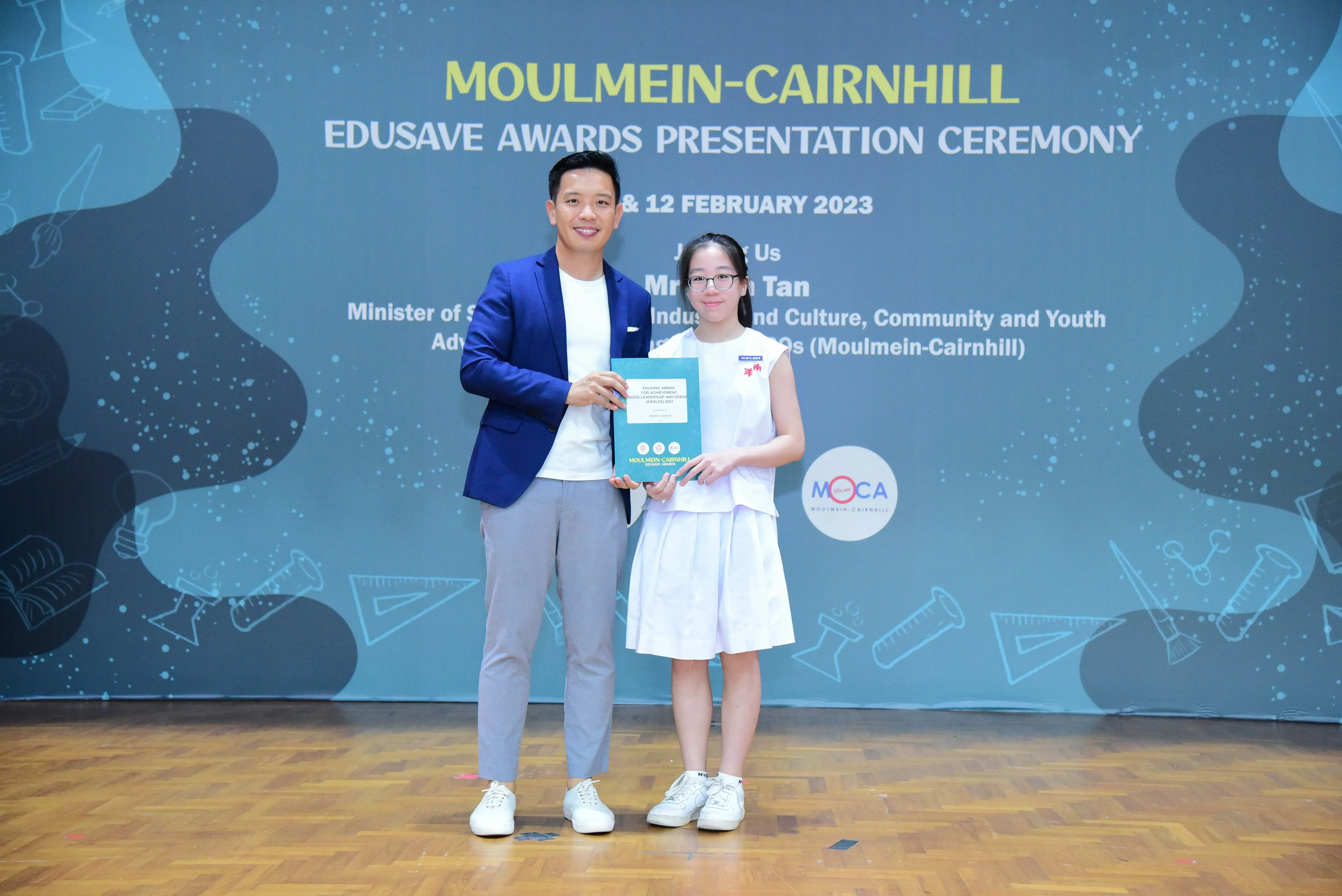 A man in a blue blazer and a young girl in white hold a certificate at the Moulmein-Cairnhill Edusave Awards Ceremony 2023.
