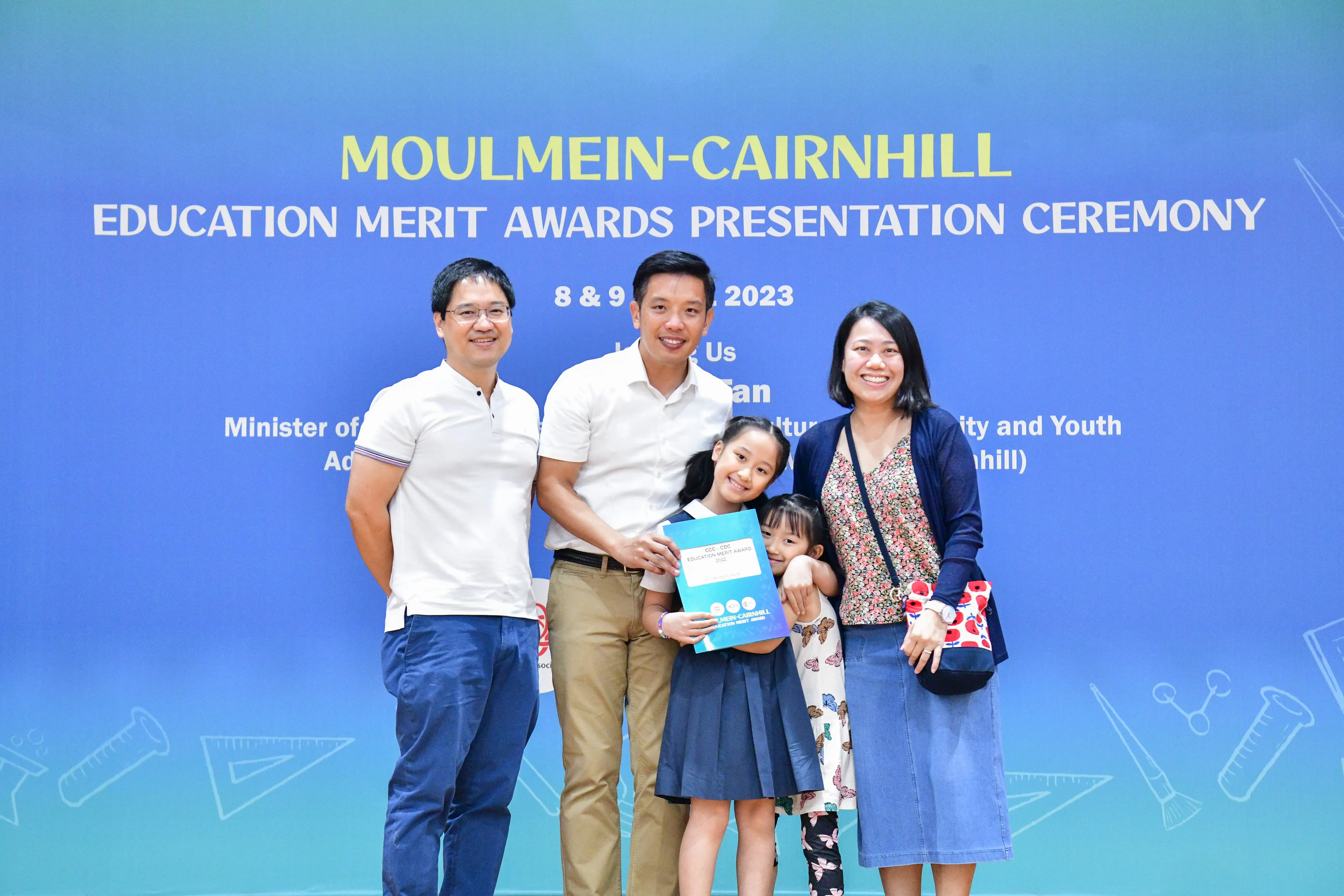 Two men, a woman, and two girls pose smiling before a Moulmein-Cairnhill Education Merit Awards banner in 2023.