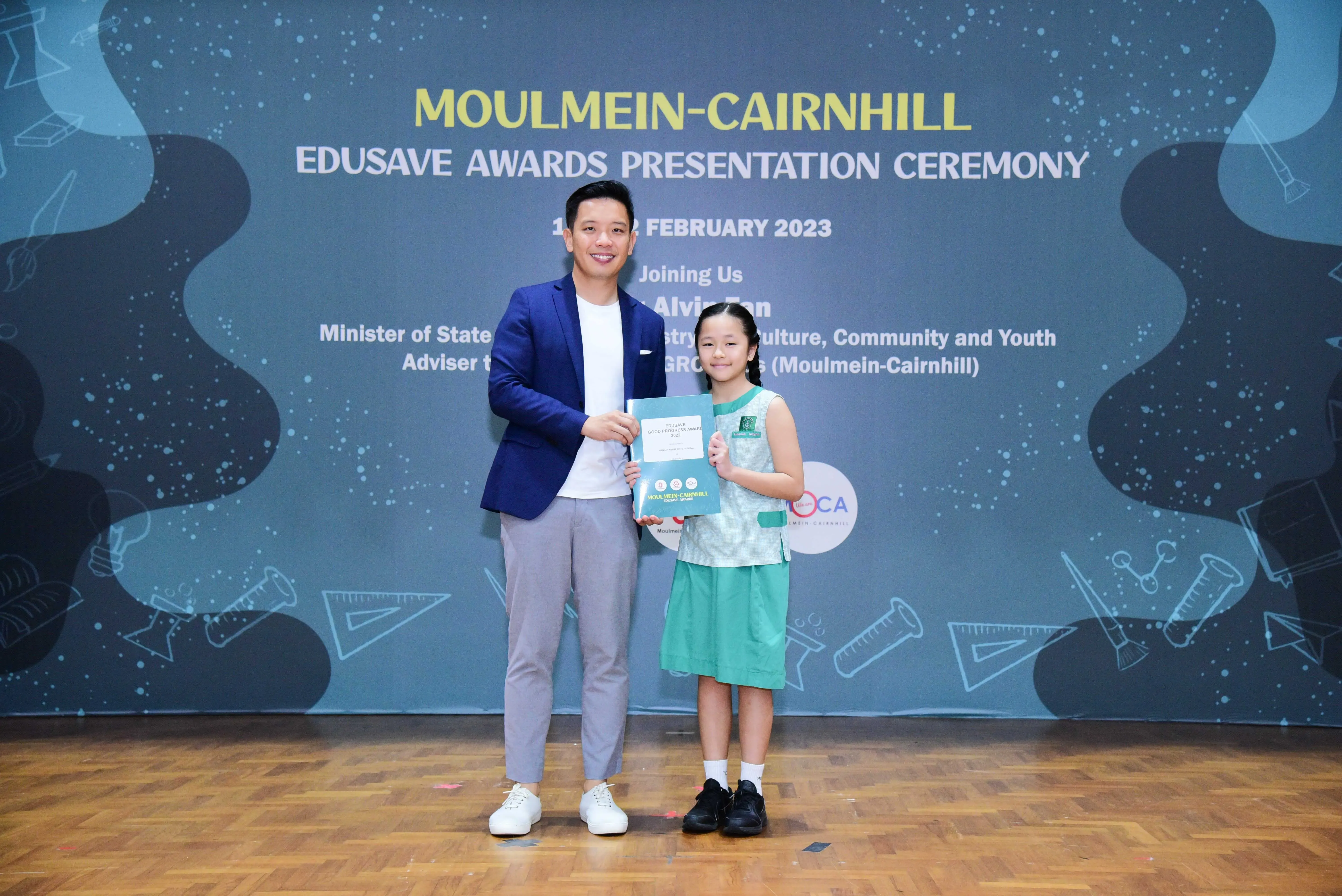 A man in a blue blazer and a young girl in a school uniform hold a certificate at the Moulmein-Cairnhill Edusave Awards 2023.
