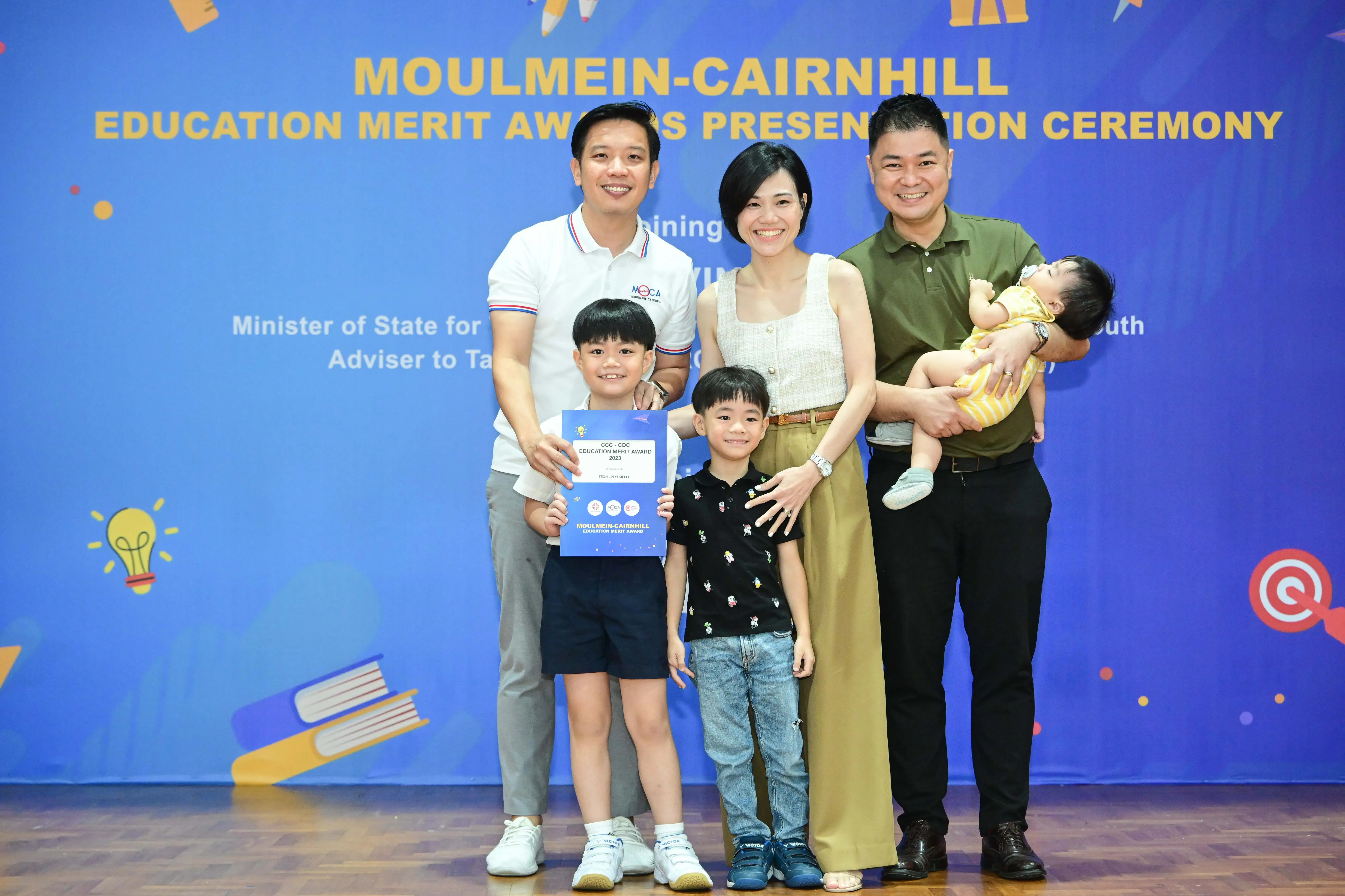 Family of five posing with an education merit award certificate at the Moulmein-Cairnhill ceremony backdrop.