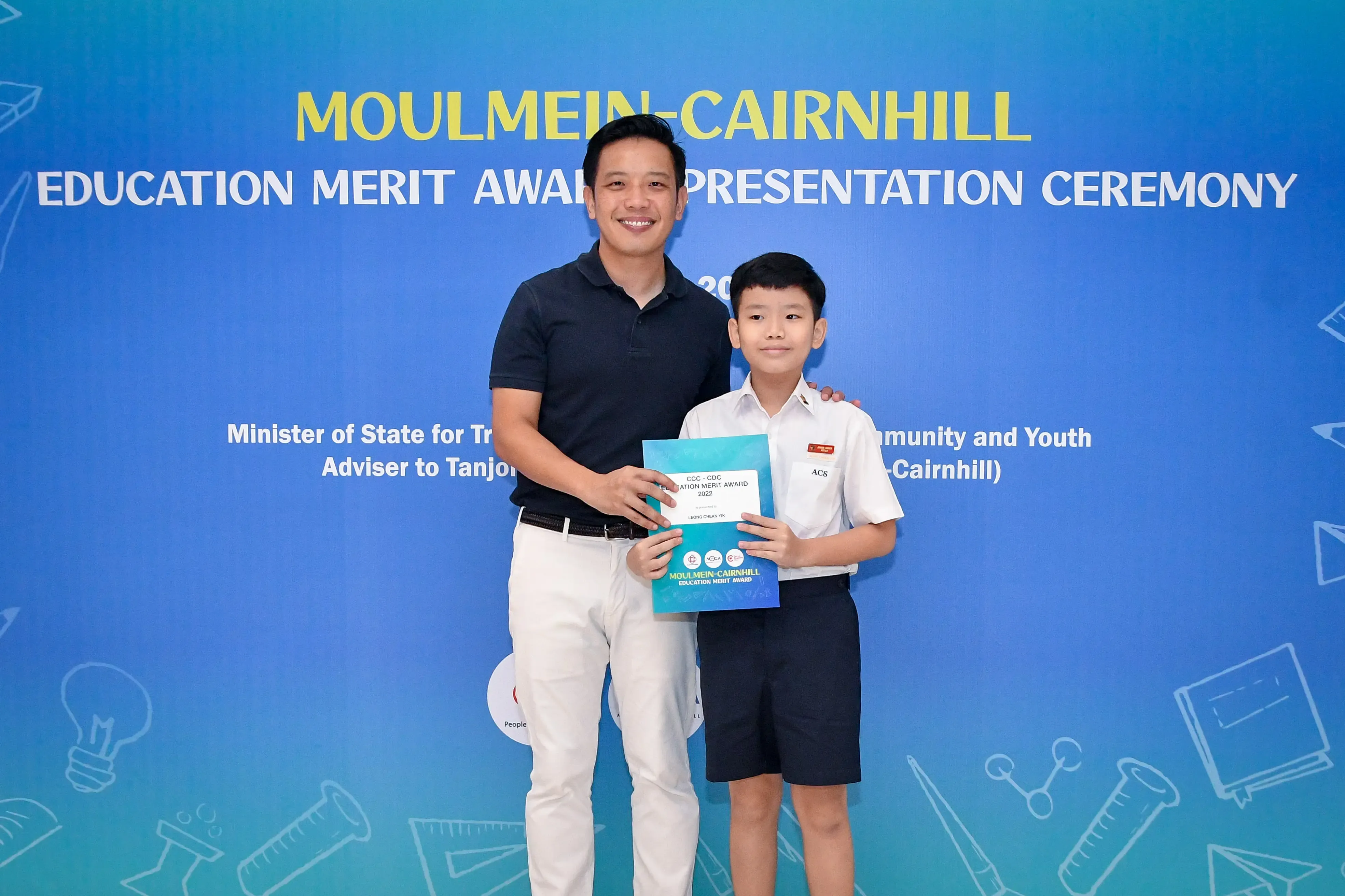 Adult man and young boy holding an award certificate at the Moulmein-Cairnhill Education Merit Award ceremony.