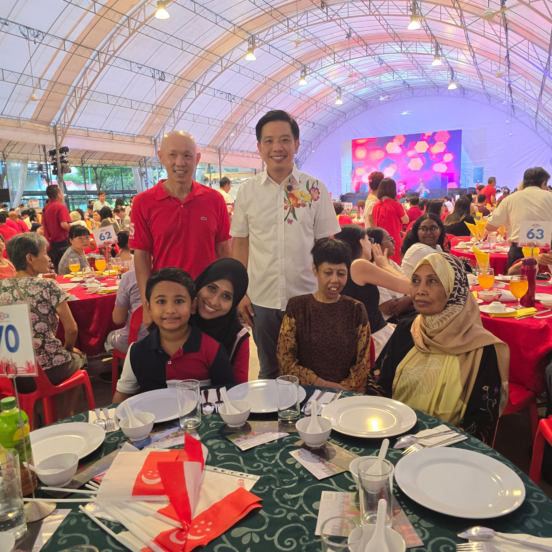 Group of people posing at a banquet table with Singapore flags, inside a large tent with a lit stage in the background.