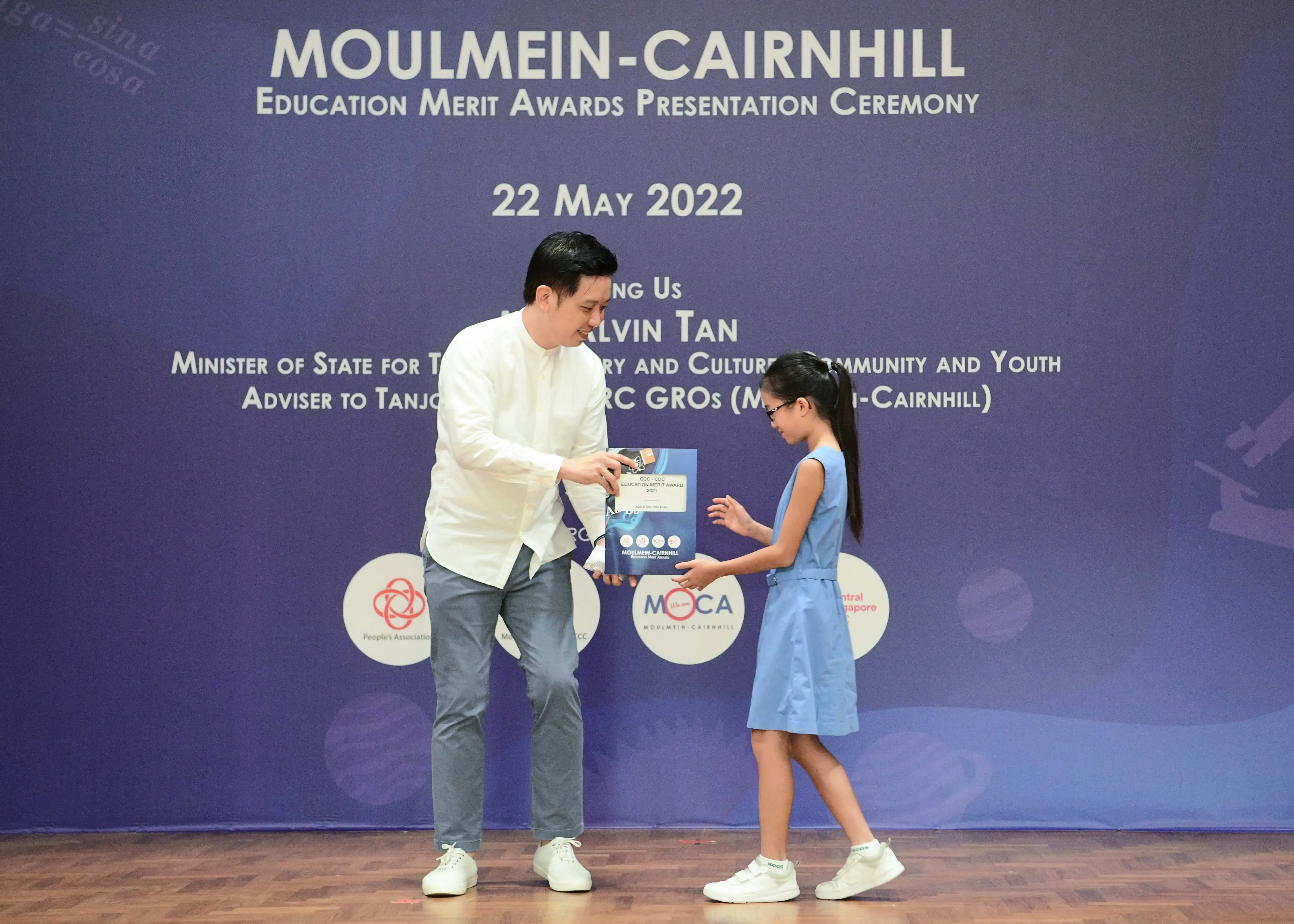 A man in white hands an award to a young girl at the Moulmein-Cairnhill Education Merit Awards on 22 May 2022.
