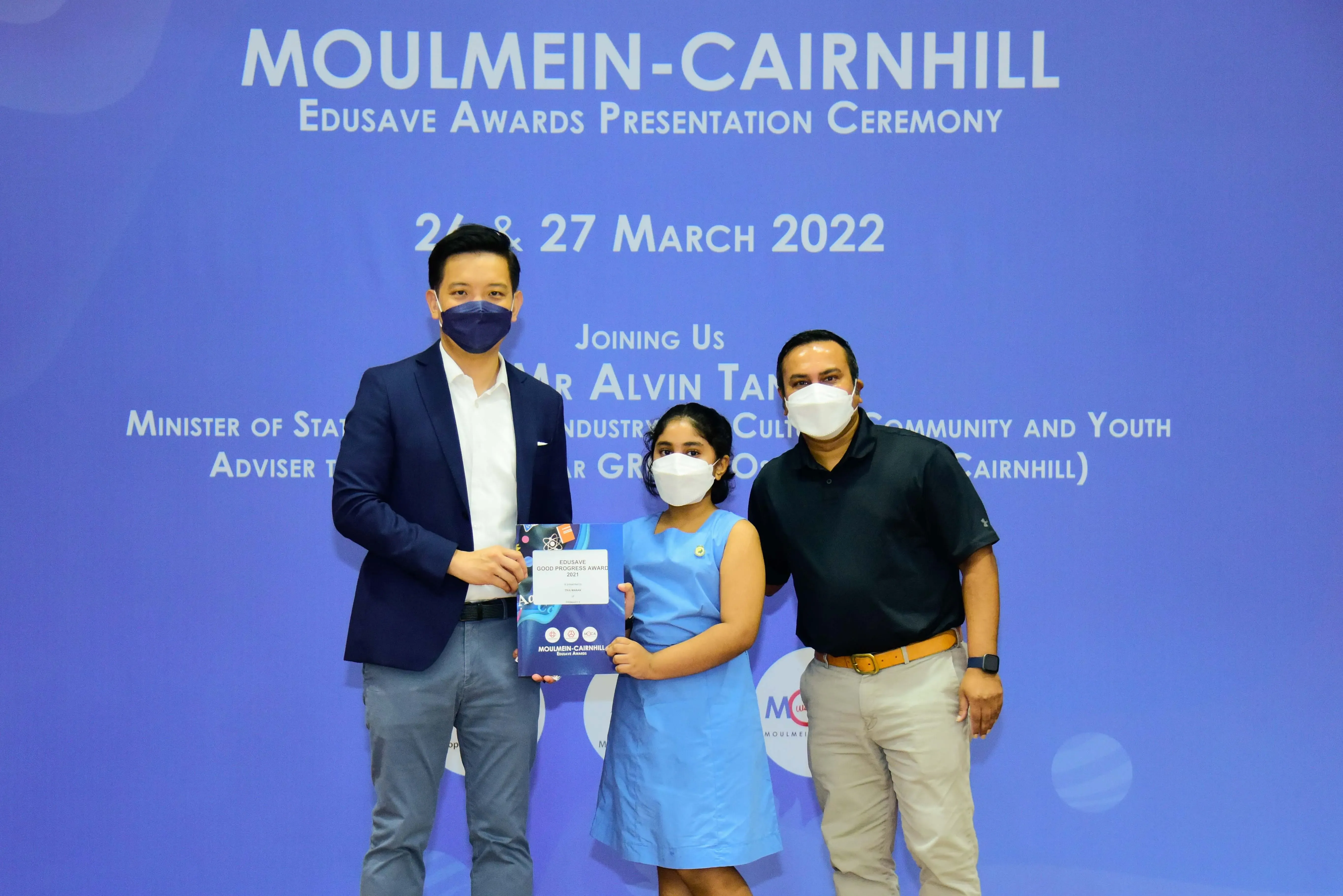 Three masked people pose at the Moulmein-Cairnhill Edusave Awards Ceremony on 26-27 March 2022, holding a certificate.