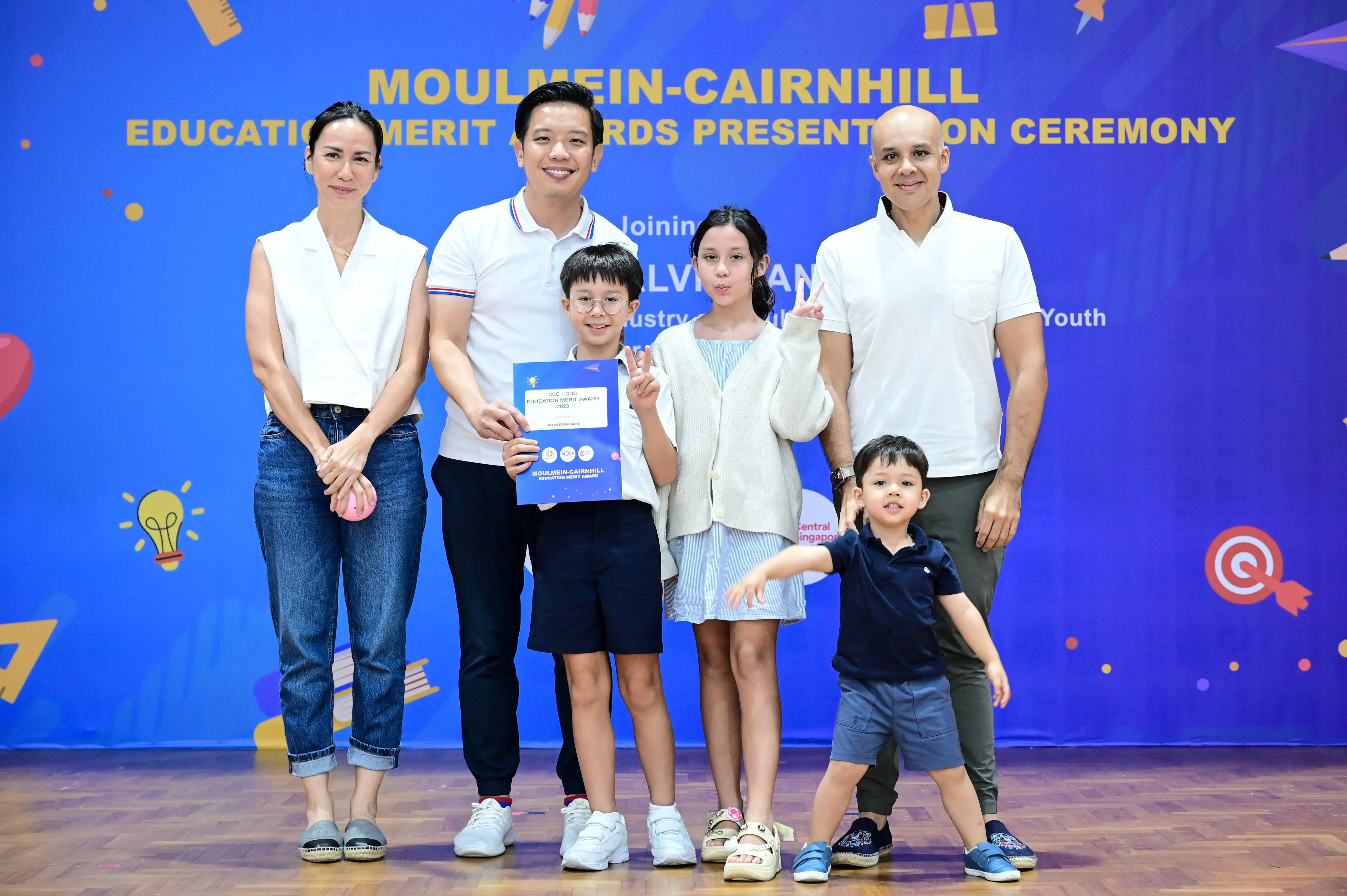 Three adults and three children pose at the Moulmein-Cairnhill Education Merit Awards Presentation Ceremony, boy holds certificate.