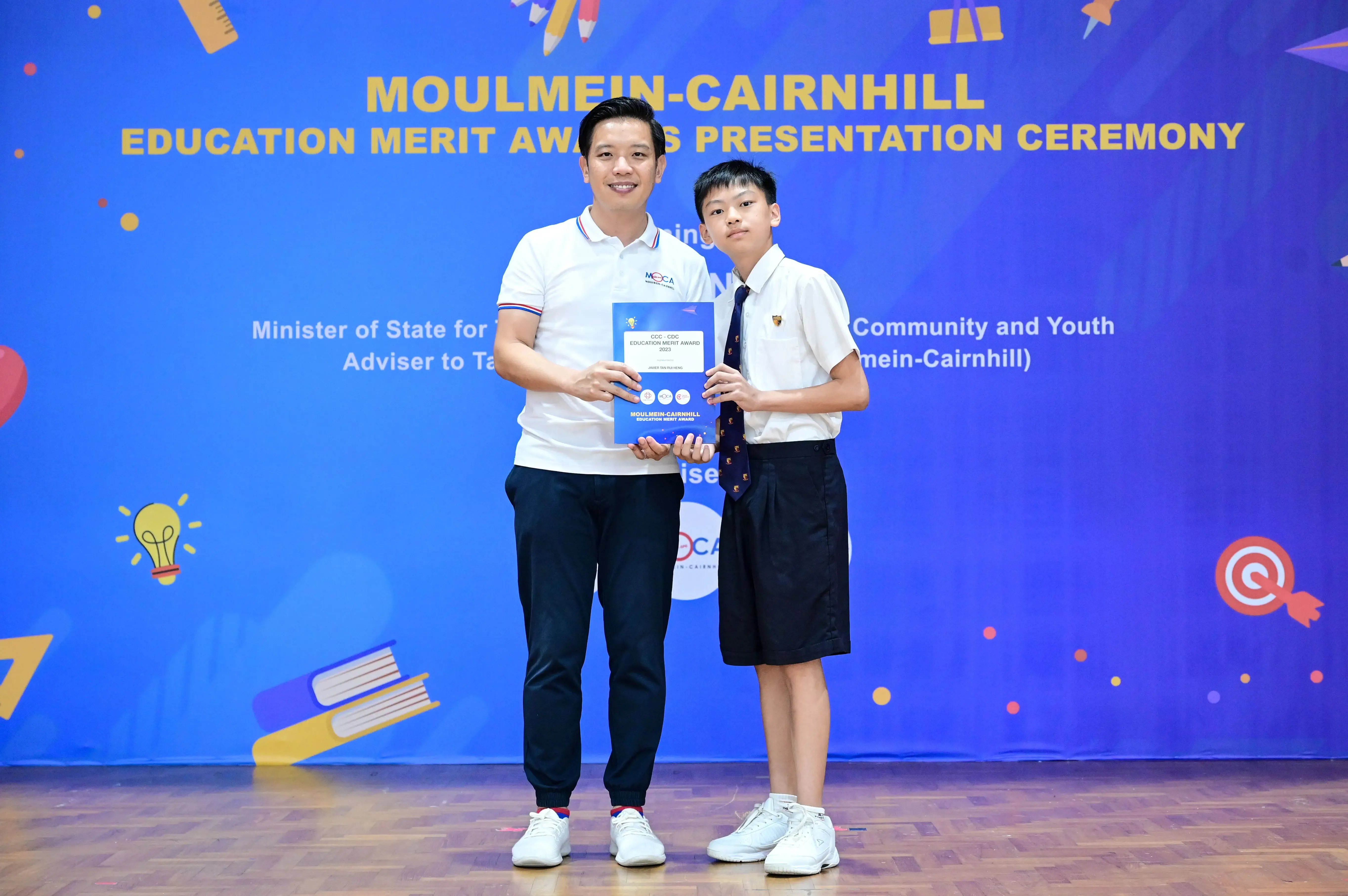 A man and a young student in uniform hold an award together at the Moulmein-Cairnhill Education Merit Awards ceremony.