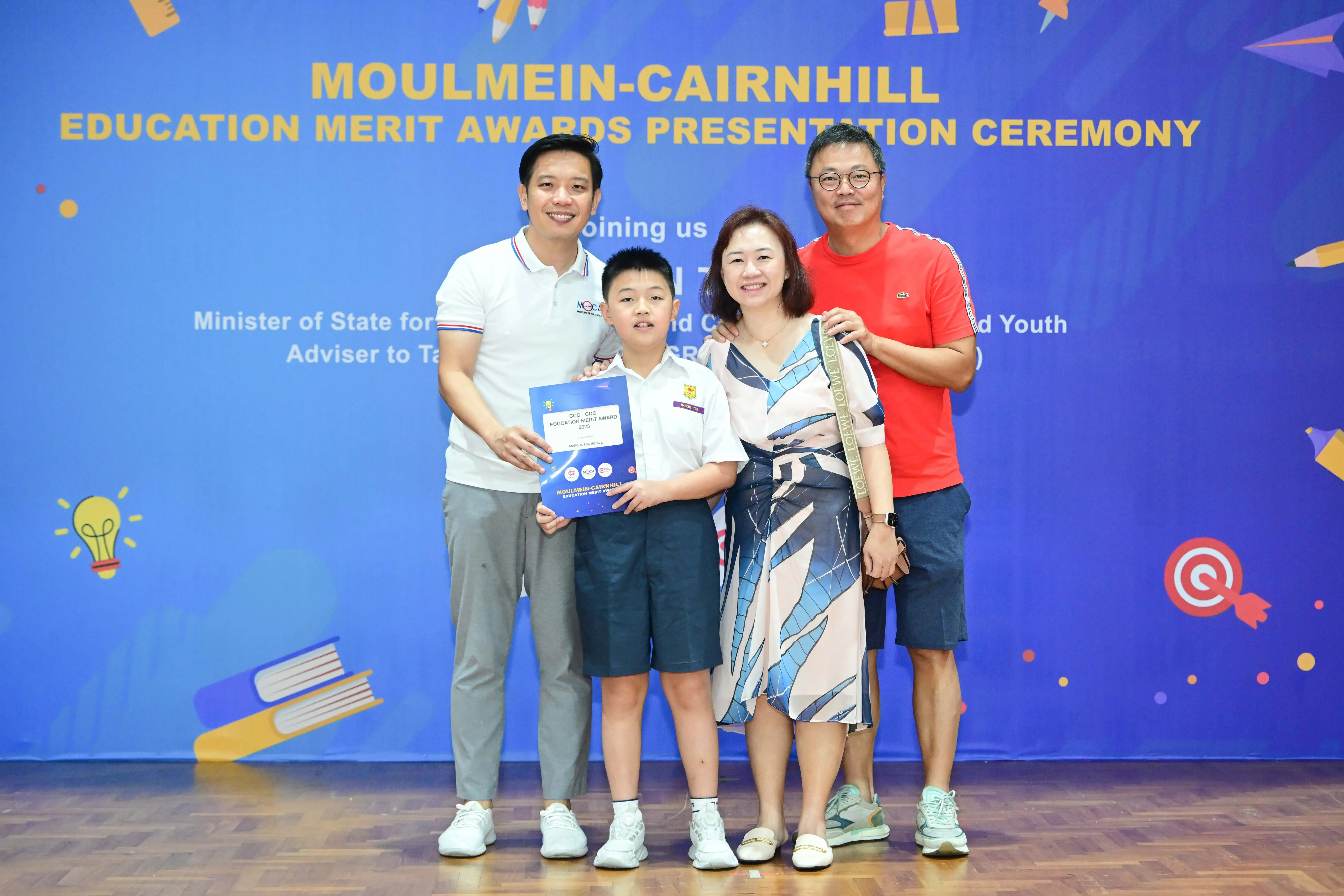 Four people pose at the Moulmein-Cairnhill Education Merit Awards ceremony, with a boy holding an award certificate.