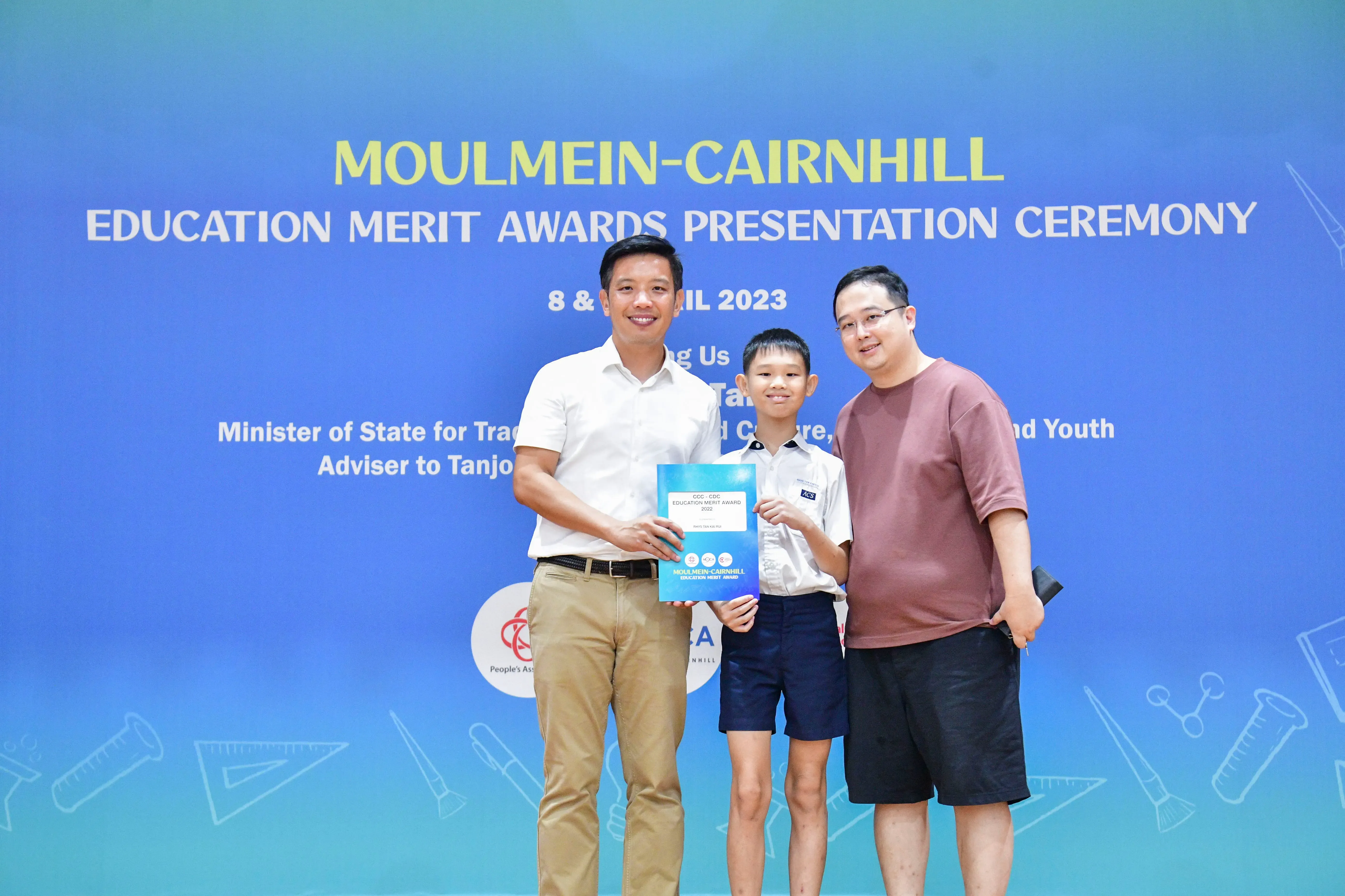 Two men and a boy holding an award certificate at the Moulmein-Cairnhill Education Merit Awards Ceremony 2023.