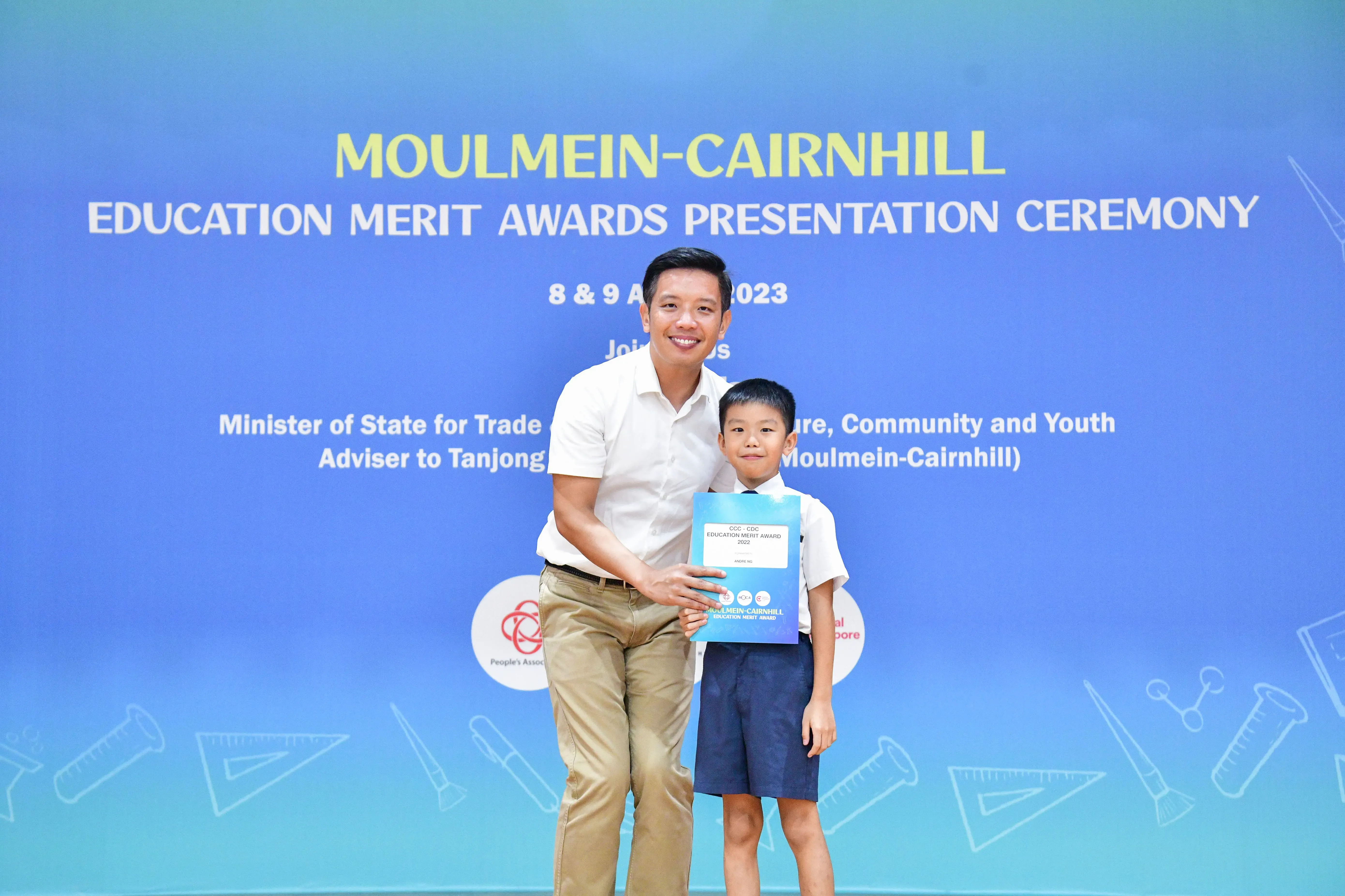 A man and young boy hold a certificate in front of a Moulmein-Cairnhill Education Merit Awards 2023 banner.
