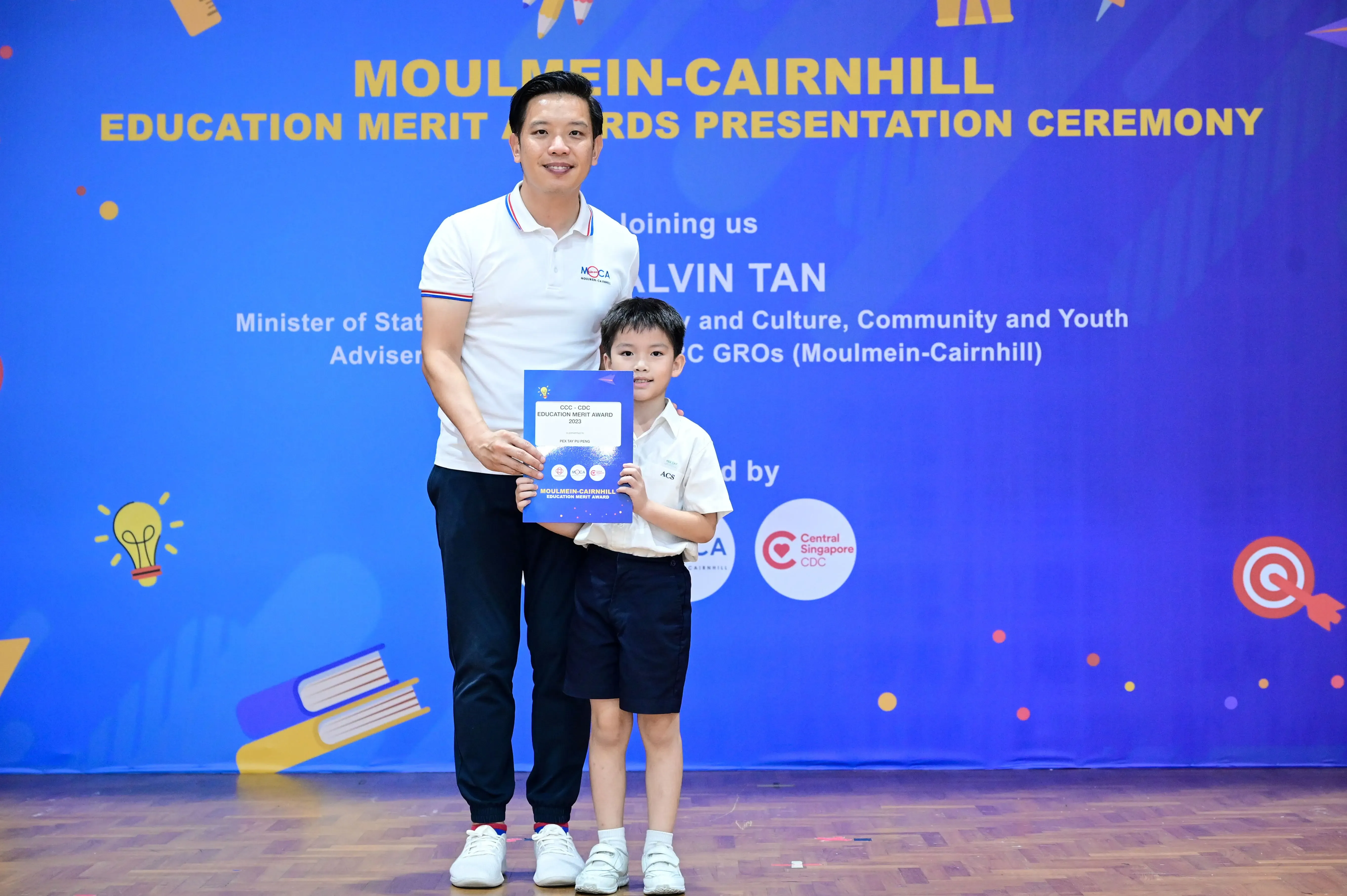 A man and young boy hold a blue certificate in front of a Moulmein-Cairnhill Education Merit Awards banner.