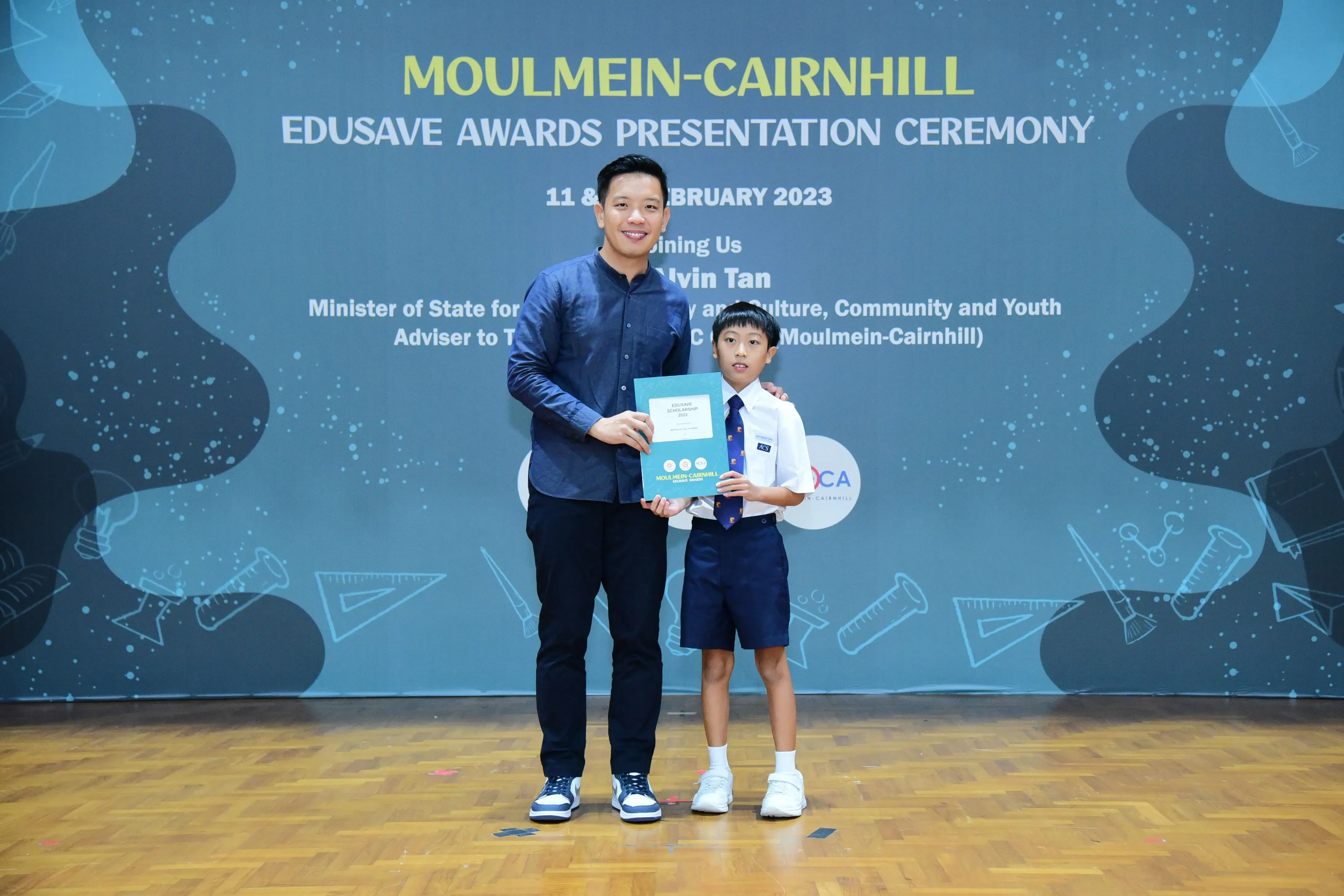 A man and a young boy in school uniform hold a certificate at the Moulmein-Cairnhill Edusave Awards Ceremony 2023.