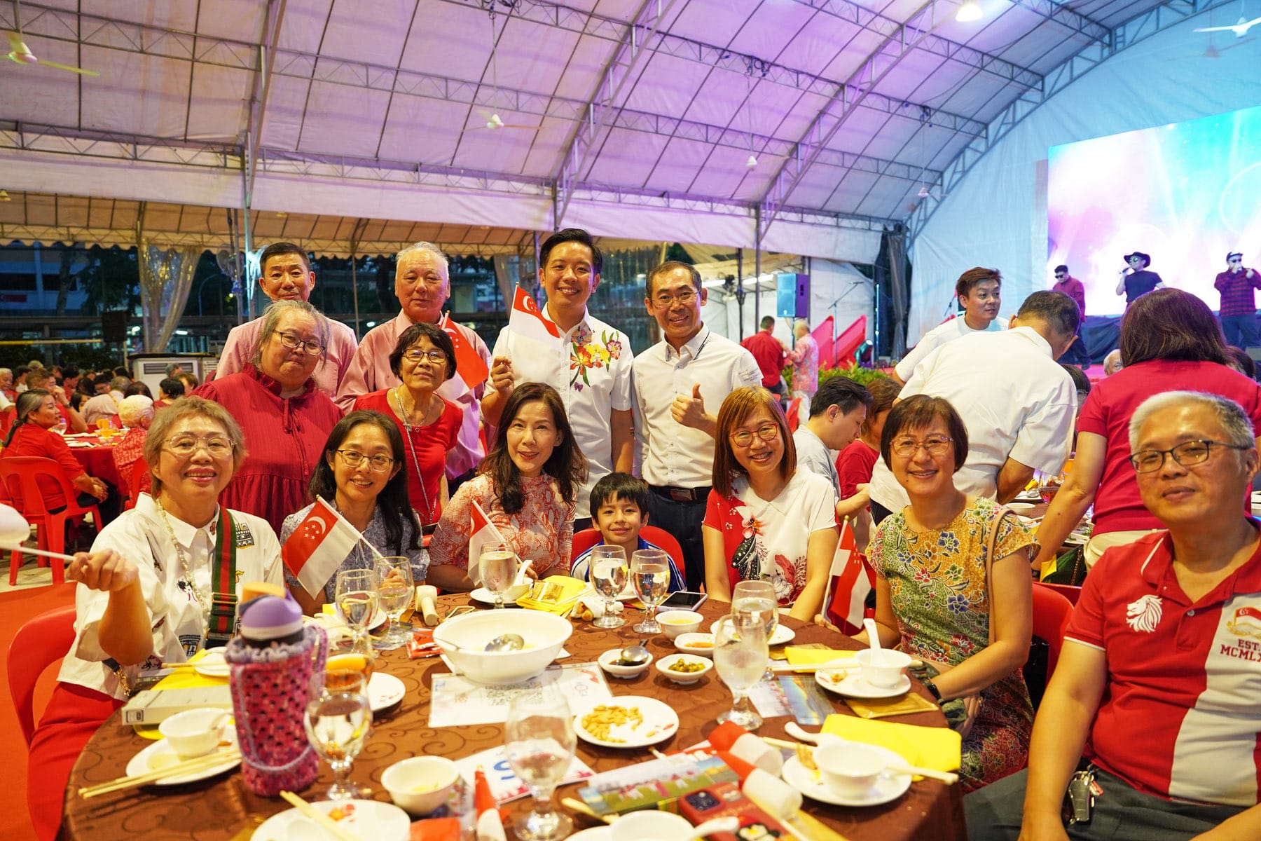 A group of smiling people at a festive dinner under a tent, holding small Singapore flags and posing for a photo.