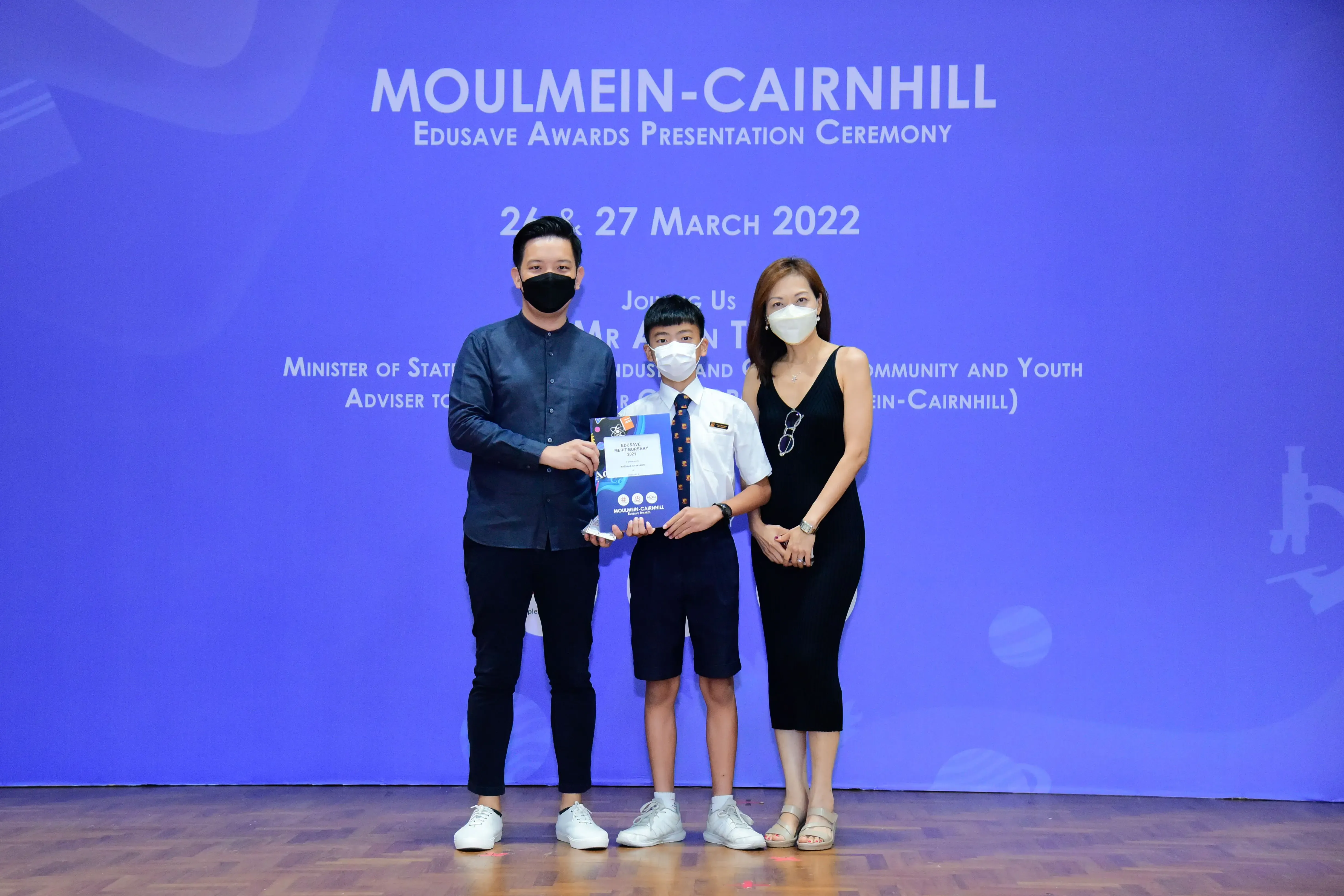 Three masked people pose at the Moulmein-Cairnhill Edusave Awards Ceremony on 26-27 March 2022, holding a certificate.