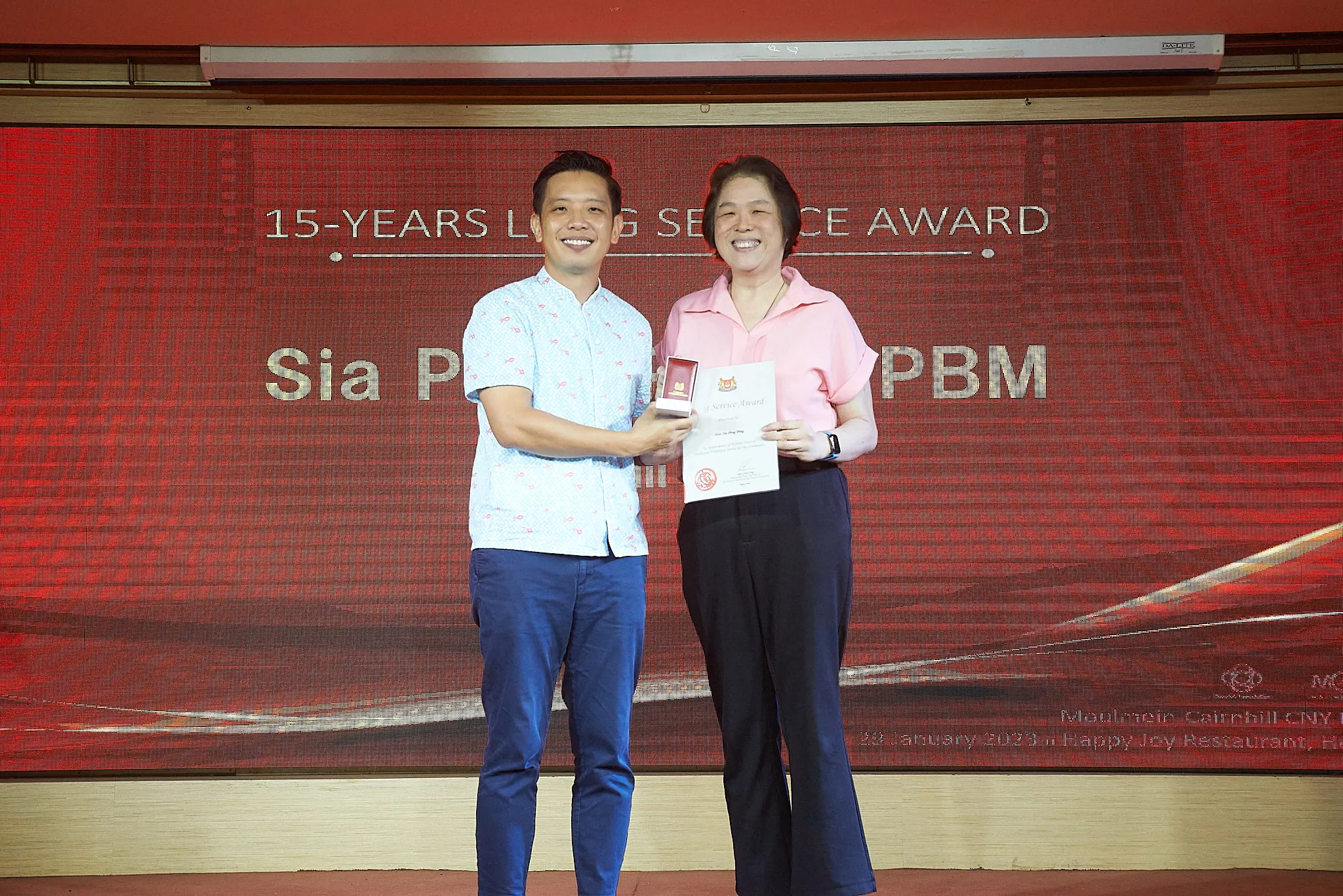 Two people smile on stage holding a certificate and award box in front of a 15-Years Long Service Award backdrop.