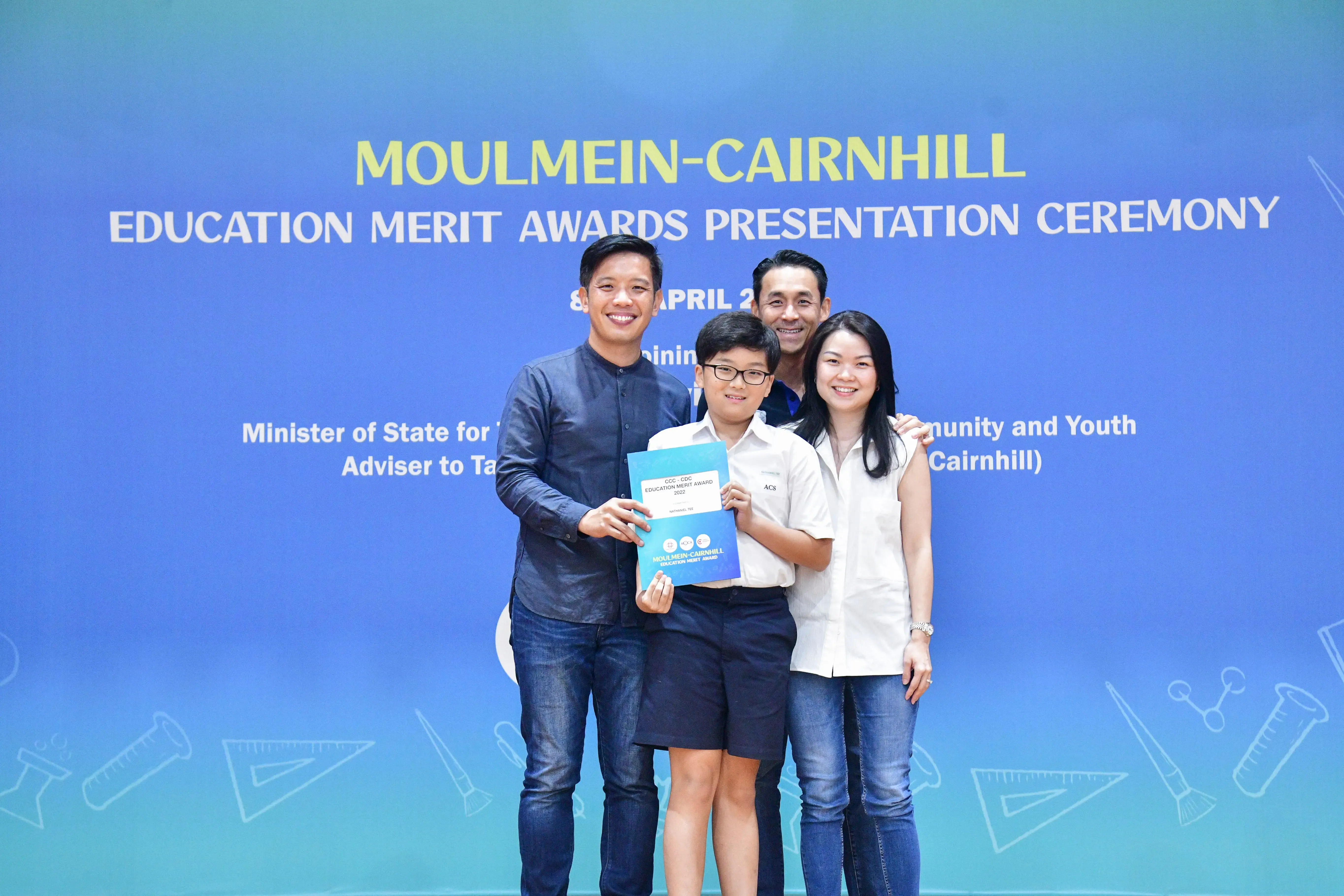 Four people pose at the Moulmein-Cairnhill Education Merit Awards Presentation Ceremony, a child holding an award folder.