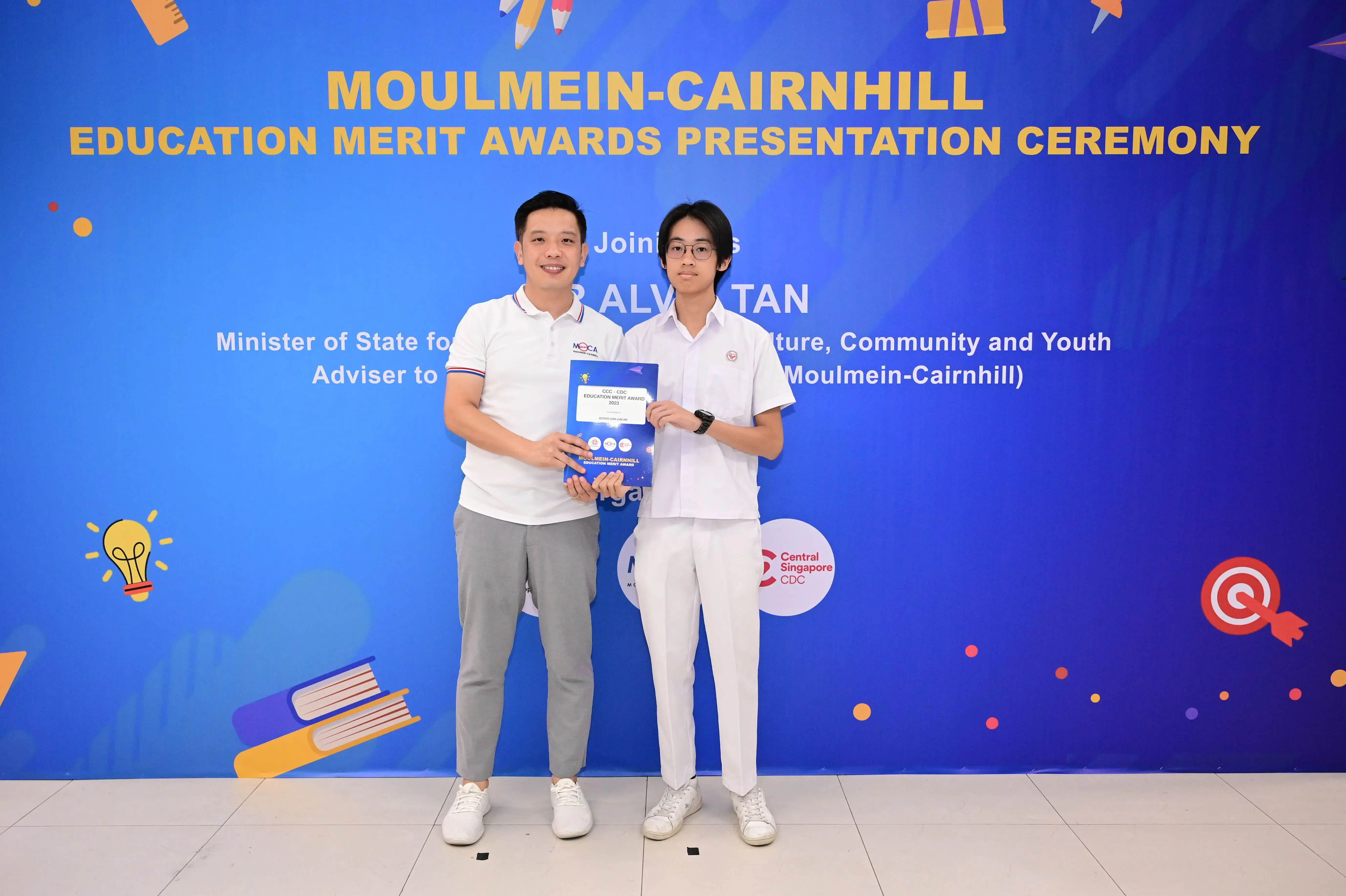 Two men hold an award booklet at the Moulmein-Cairnhill Education Merit Awards Presentation Ceremony backdrop.