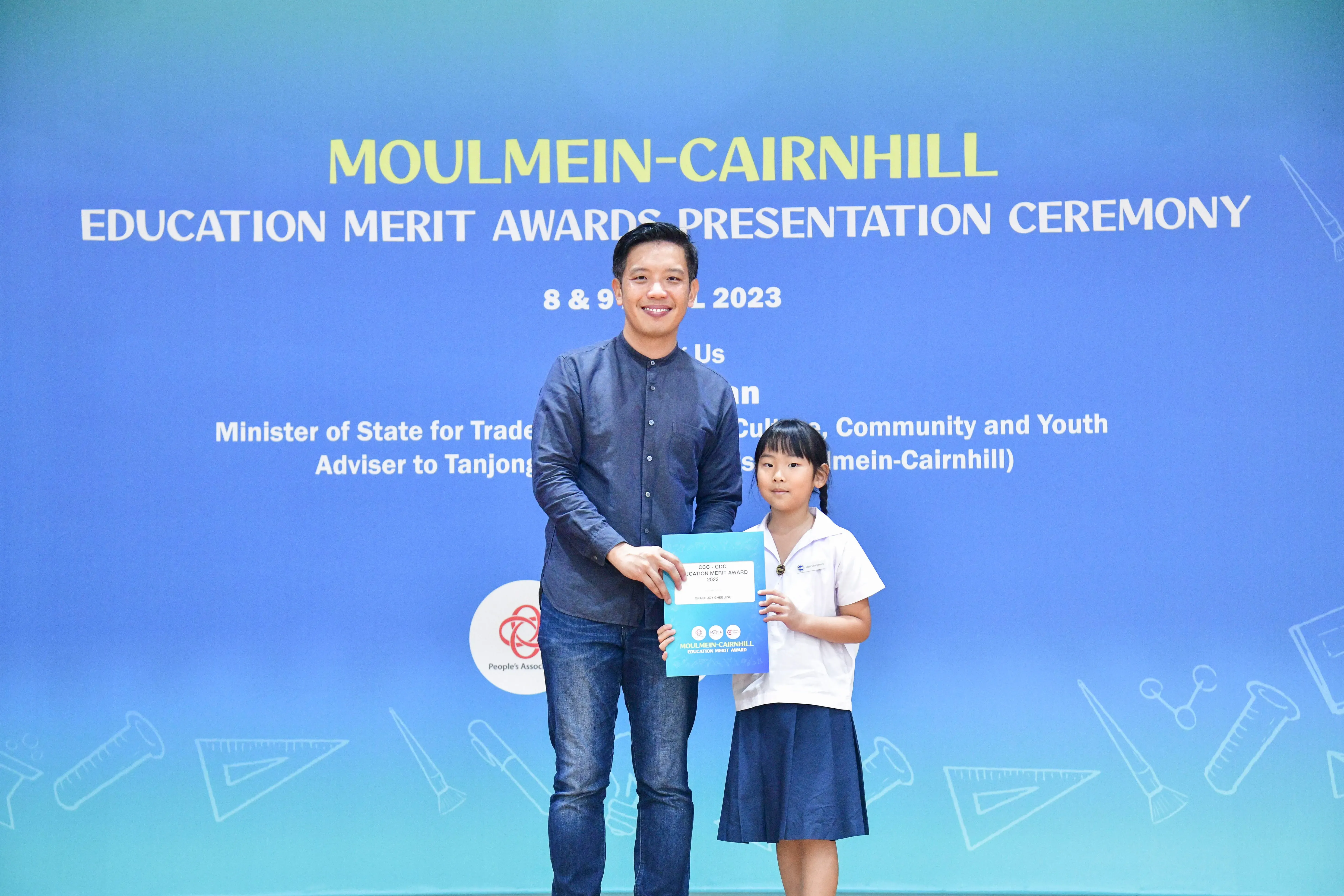 A man and a young girl hold a certificate together in front of a Moulmein-Cairnhill Education Merit Awards banner.