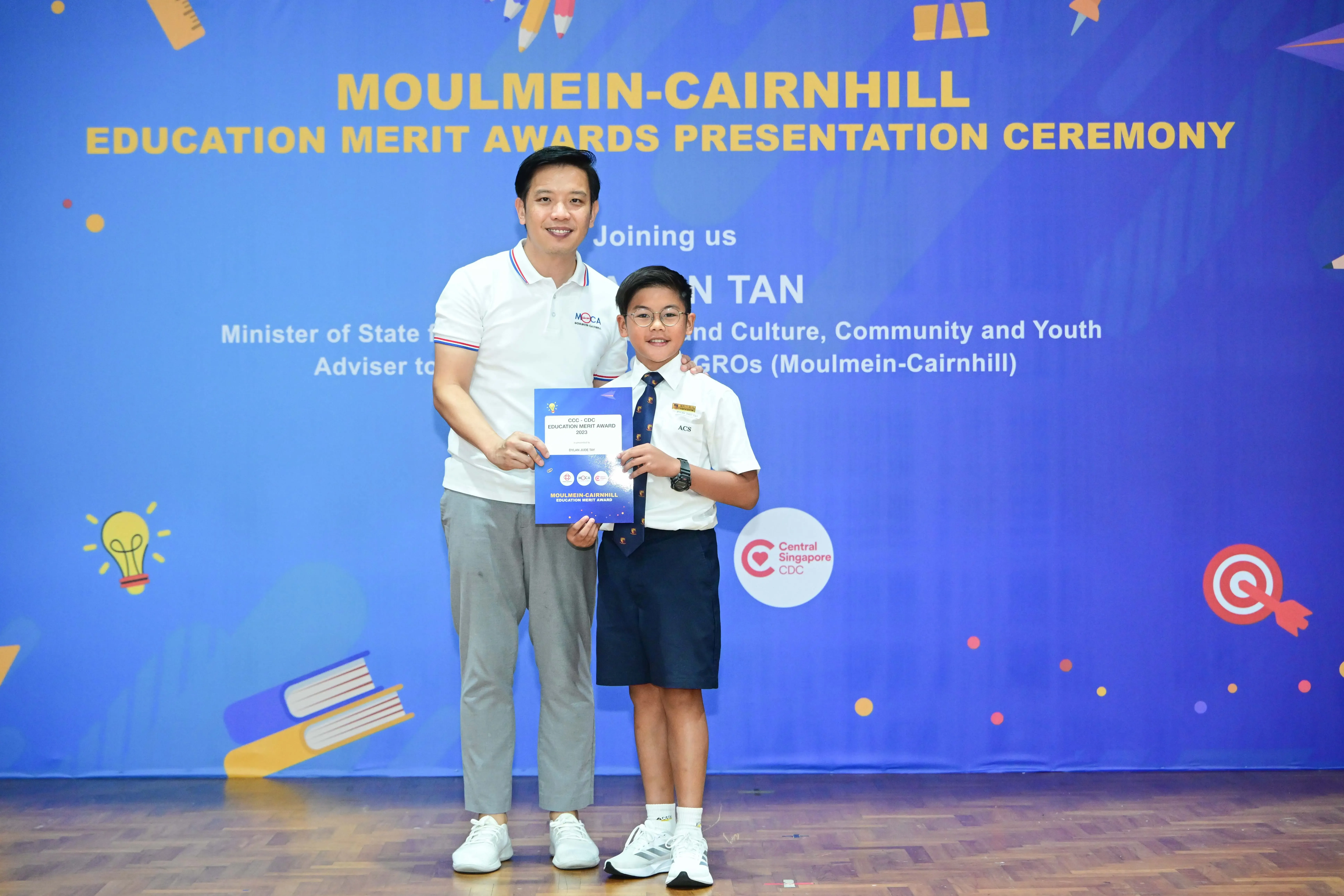 A man and a young boy in school uniform hold a certificate at the Moulmein-Cairnhill Education Merit Awards ceremony.
