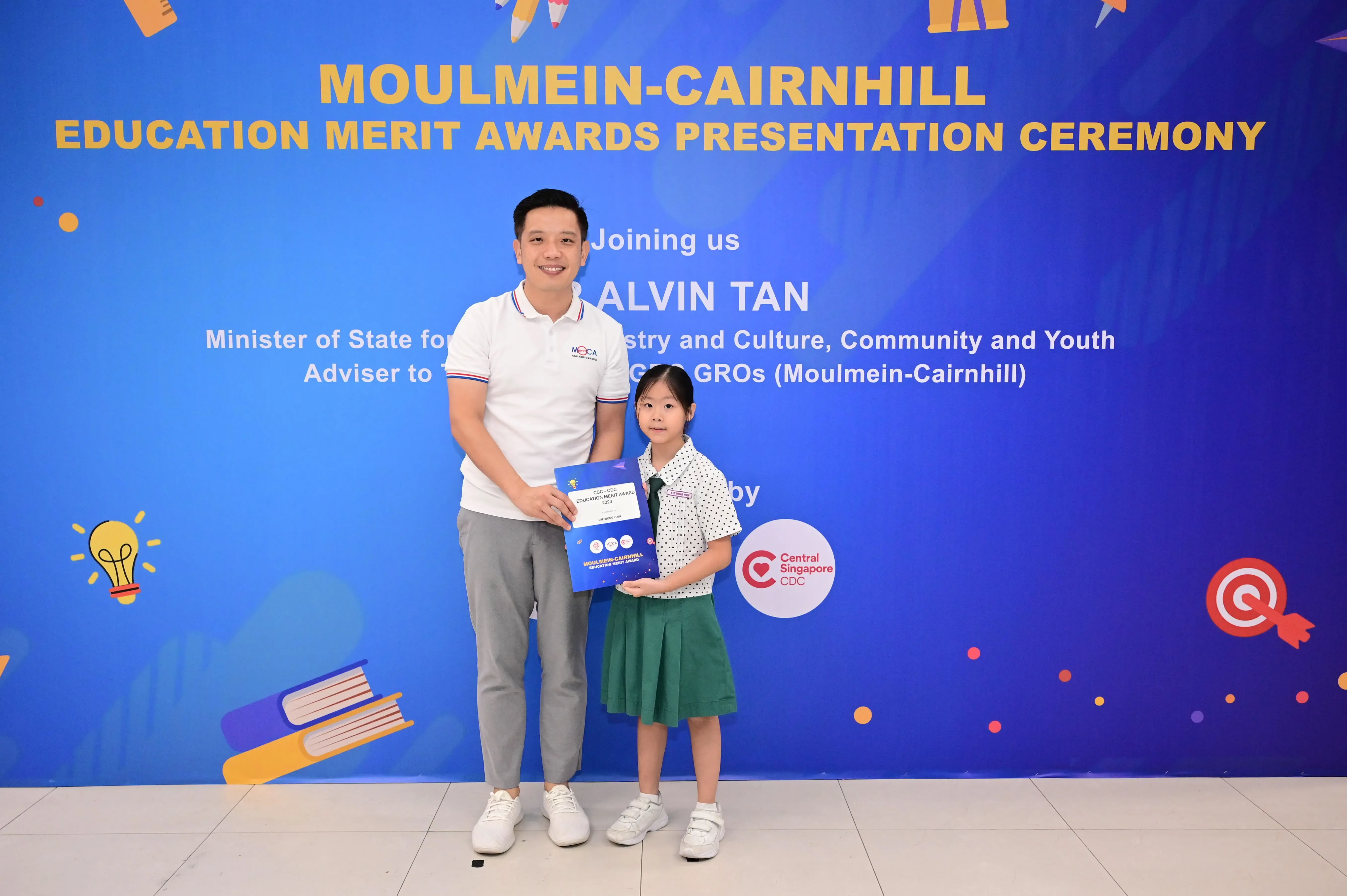 A man and a young girl hold a blue award booklet at the Moulmein-Cairnhill Education Merit Awards Ceremony.