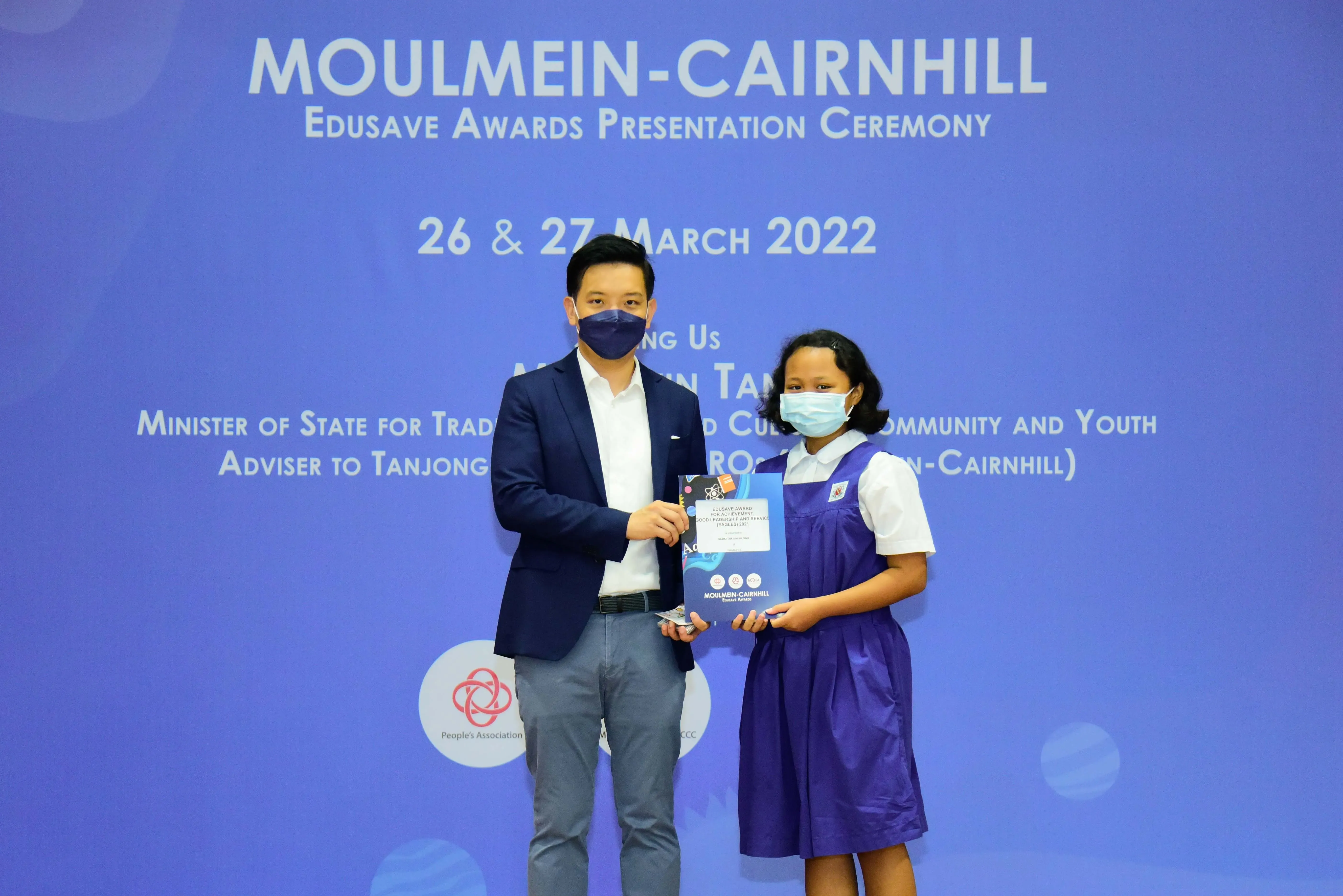 A man and a young girl in school uniform, both masked, hold an award at the Moulmein-Cairnhill Edusave ceremony.