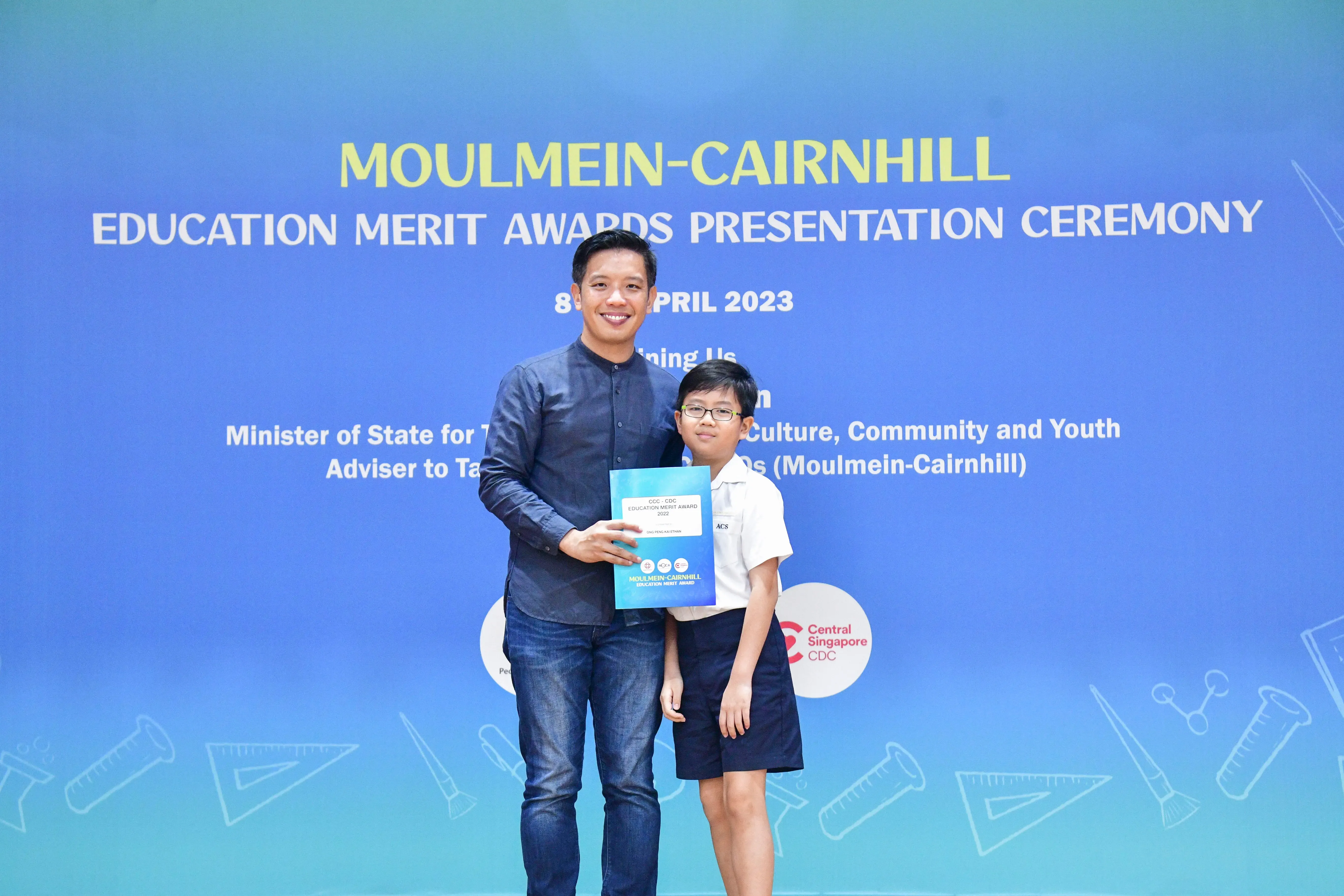 An adult and a young boy hold a certificate at the Moulmein-Cairnhill Education Merit Awards Ceremony, April 2023.