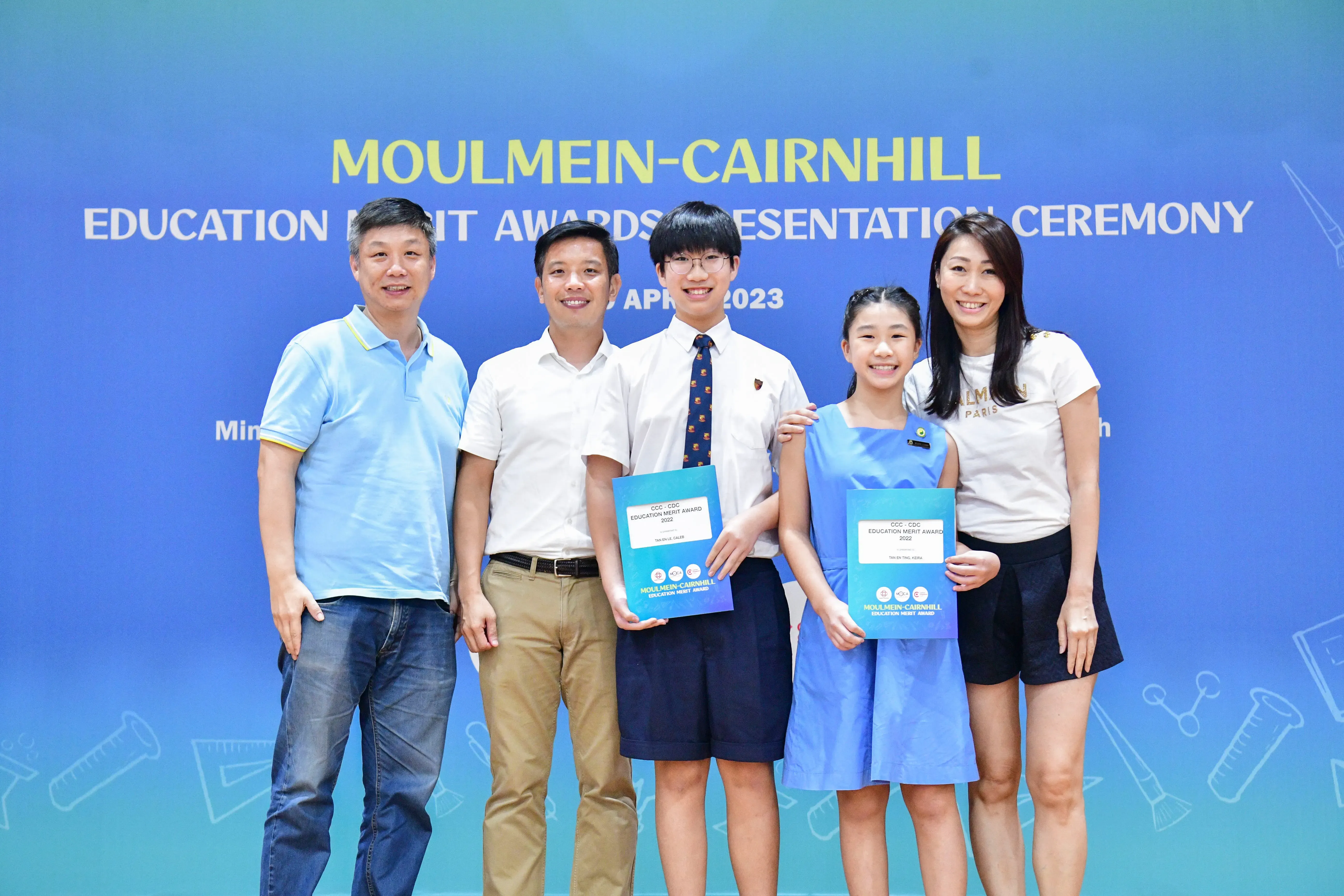 Five people pose at the Moulmein-Cairnhill Education Merit Awards Ceremony April 2023, two students holding award folders.