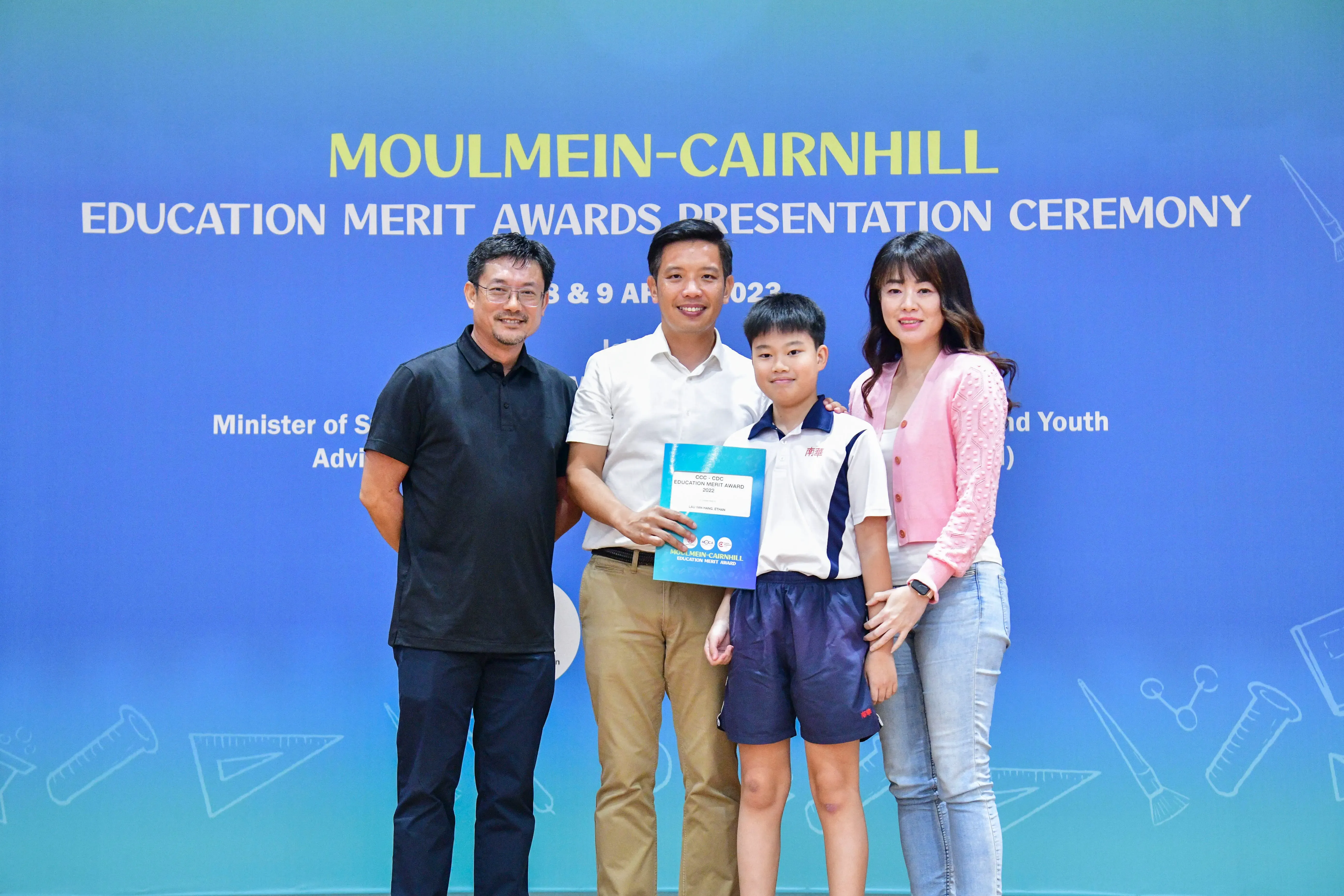 Four people pose before a Moulmein-Cairnhill Education Merit Awards Presentation Ceremony banner, a boy holding an award certificate.