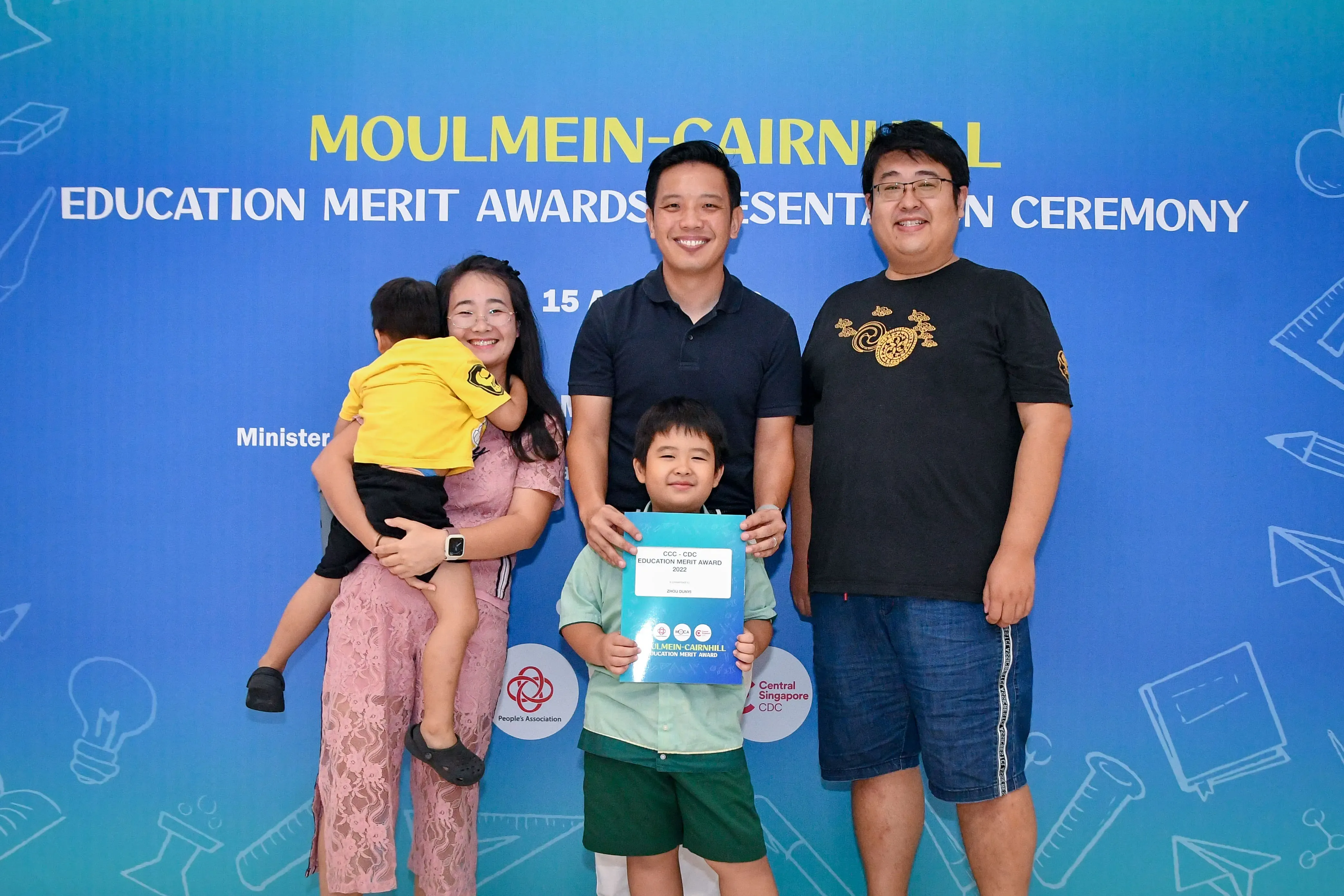 Four adults and two children pose at the Moulmein-Cairnhill Education Merit Awards ceremony, boy holding a certificate.