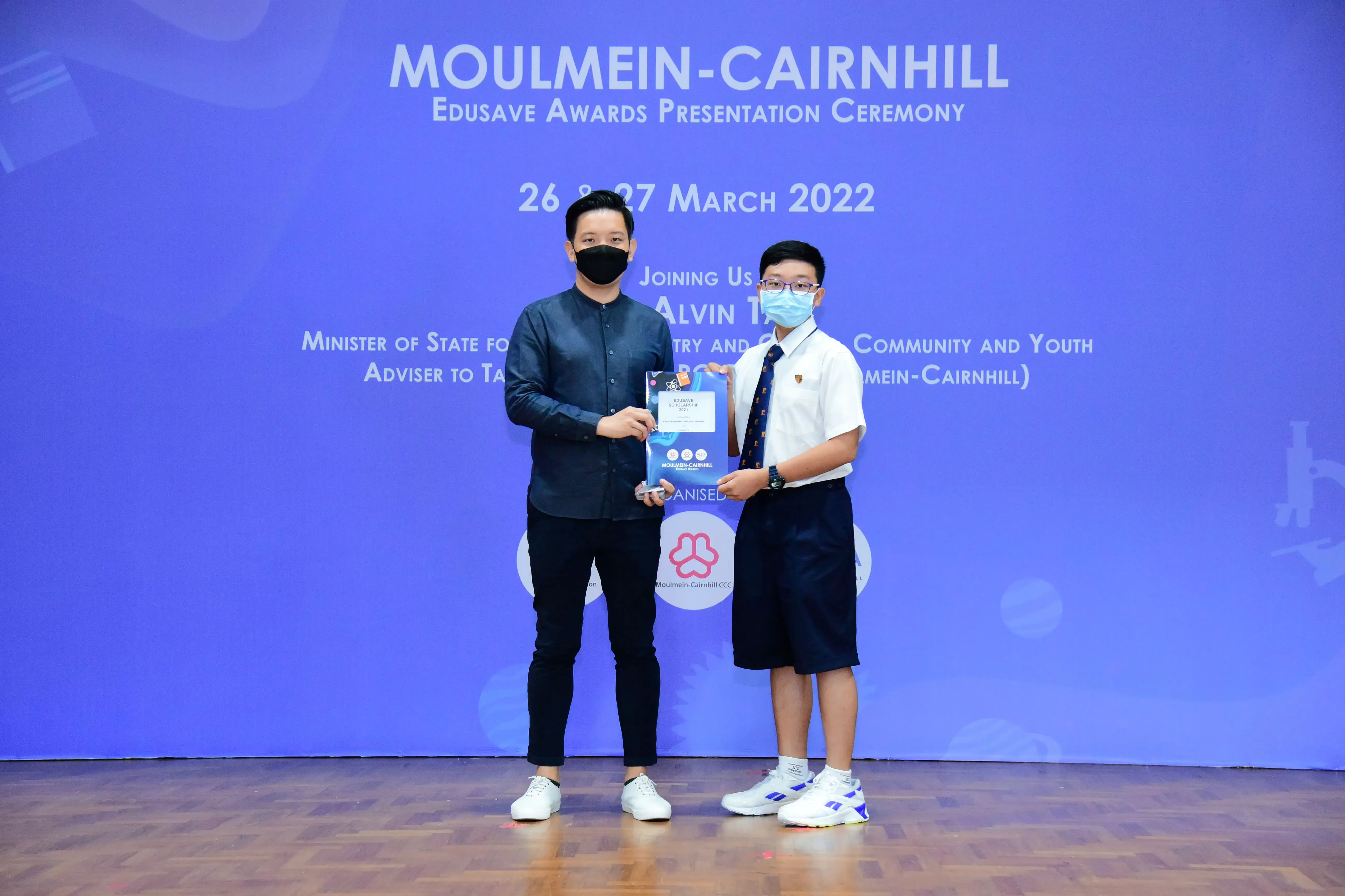 An adult and a student in uniform hold an award at the Moulmein-Cairnhill Edusave Awards Ceremony, March 2022.