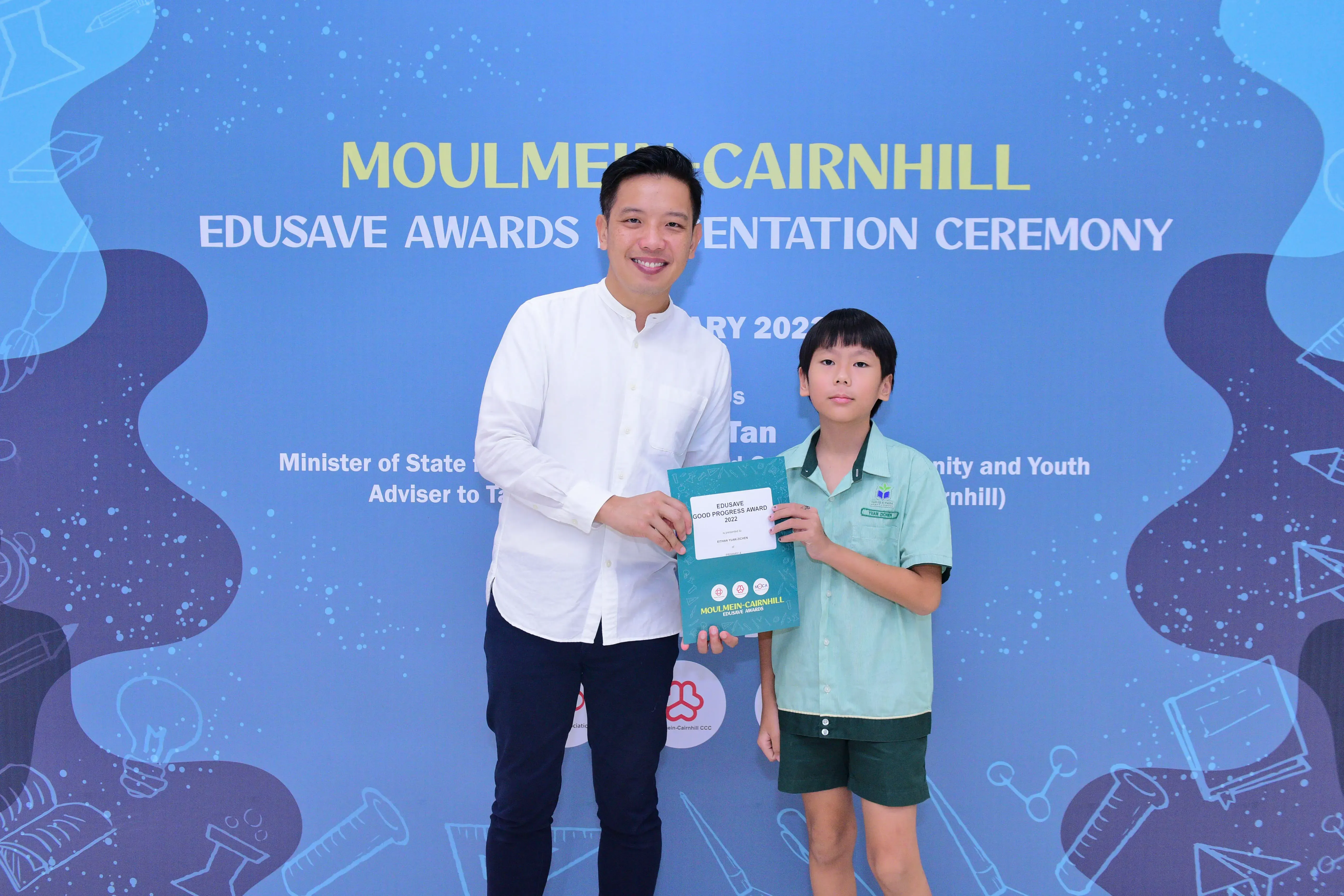 Man in white shirt and young boy in school uniform hold an Edusave award at Moulmein-Cairnhill ceremony.