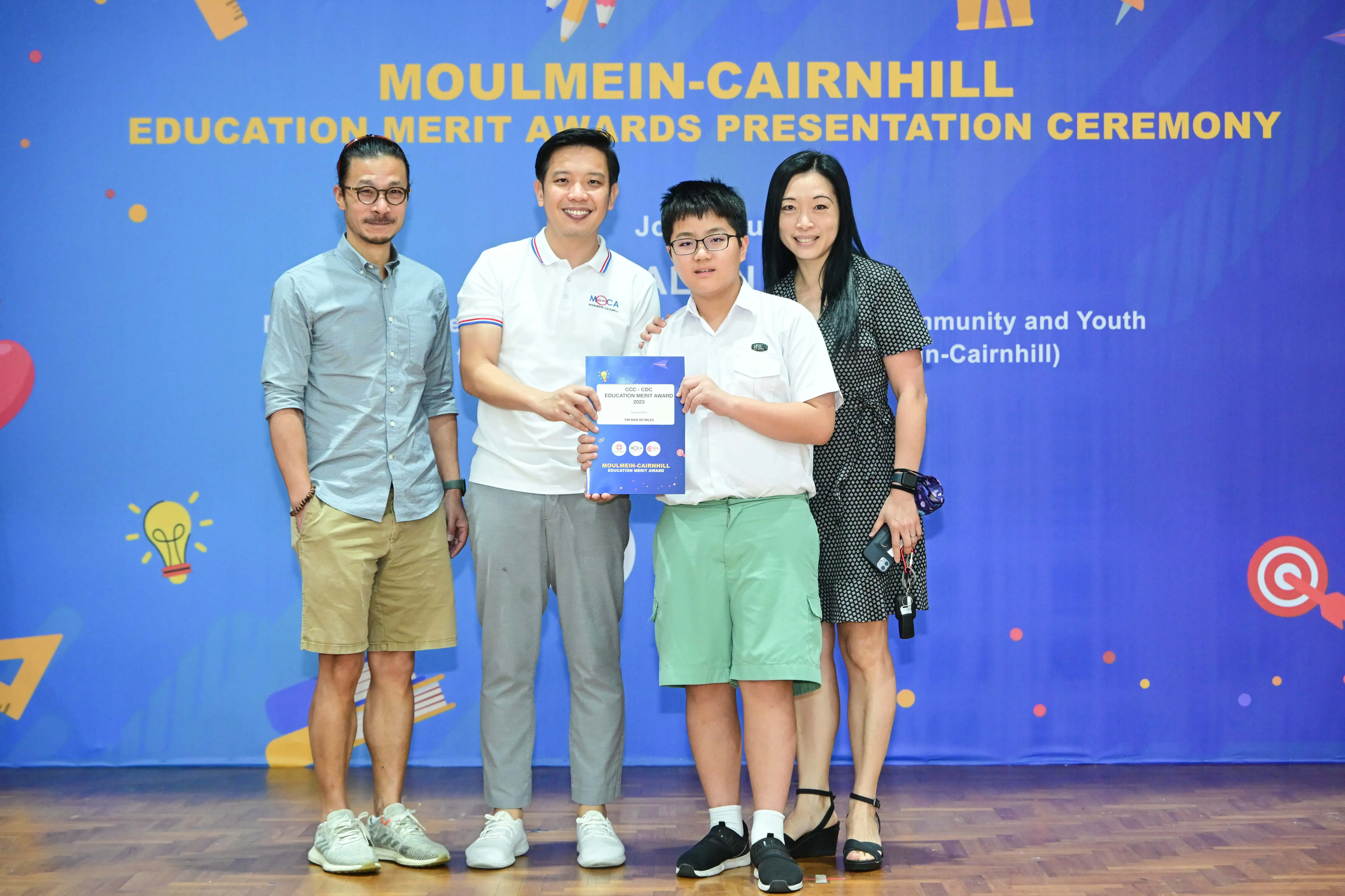 Four people pose at the Moulmein-Cairnhill Education Merit Awards ceremony, with a boy holding a blue certificate.