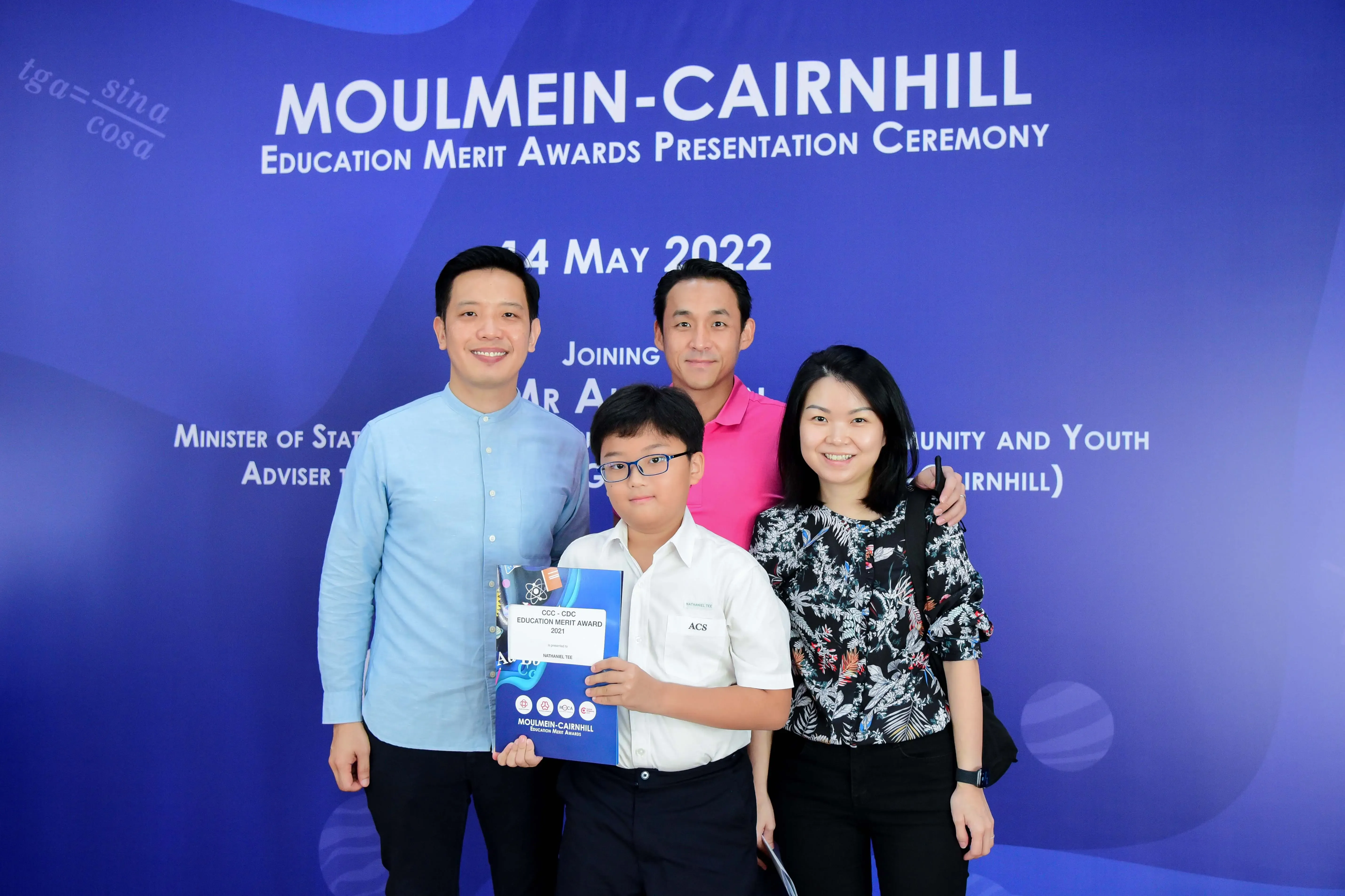 Four people pose before a Moulmein-Cairnhill Education Merit Awards Presentation Ceremony banner dated 14 May 2022.