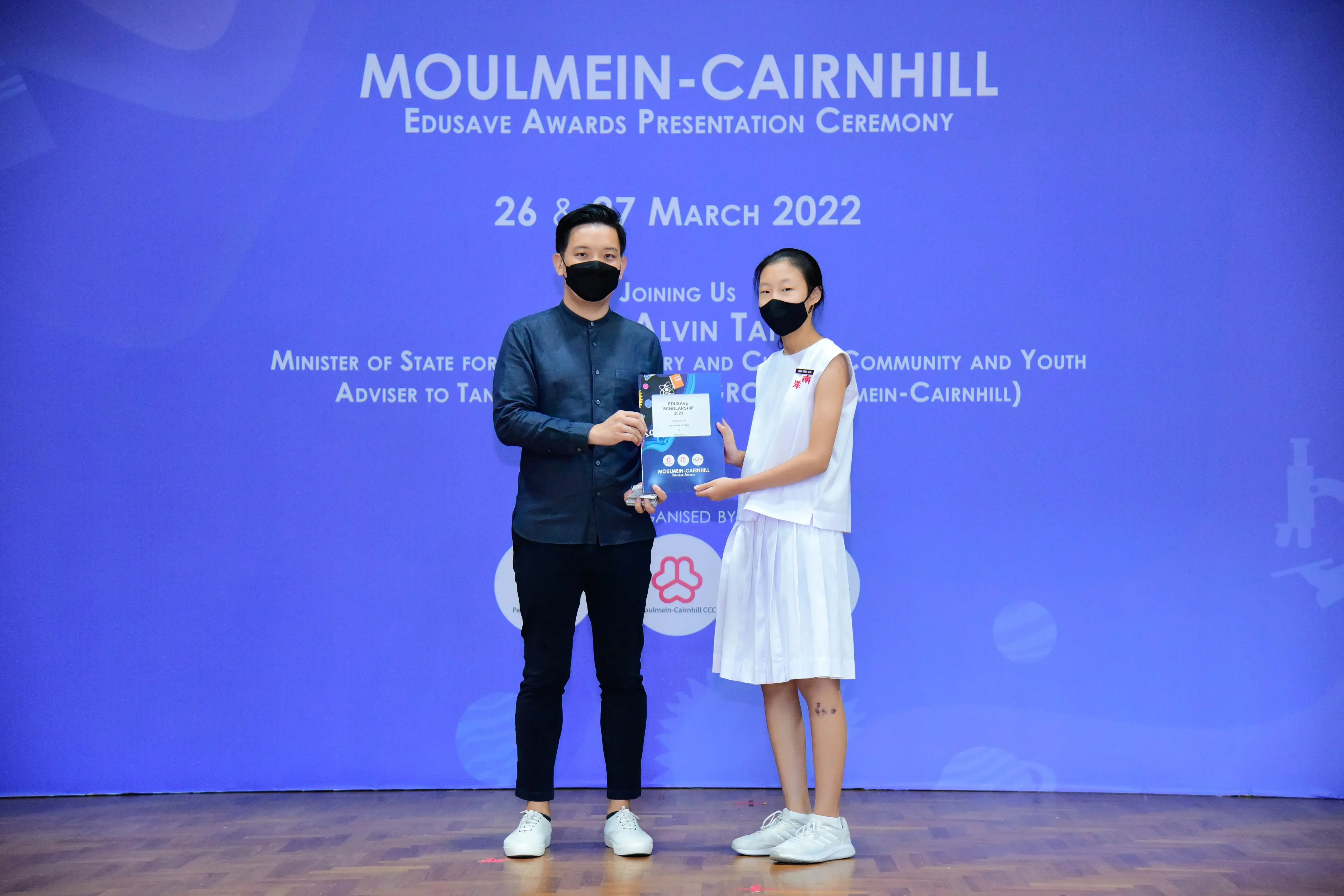 Two masked people hold an award at the Moulmein-Cairnhill Edusave Awards Presentation Ceremony, March 2022.
