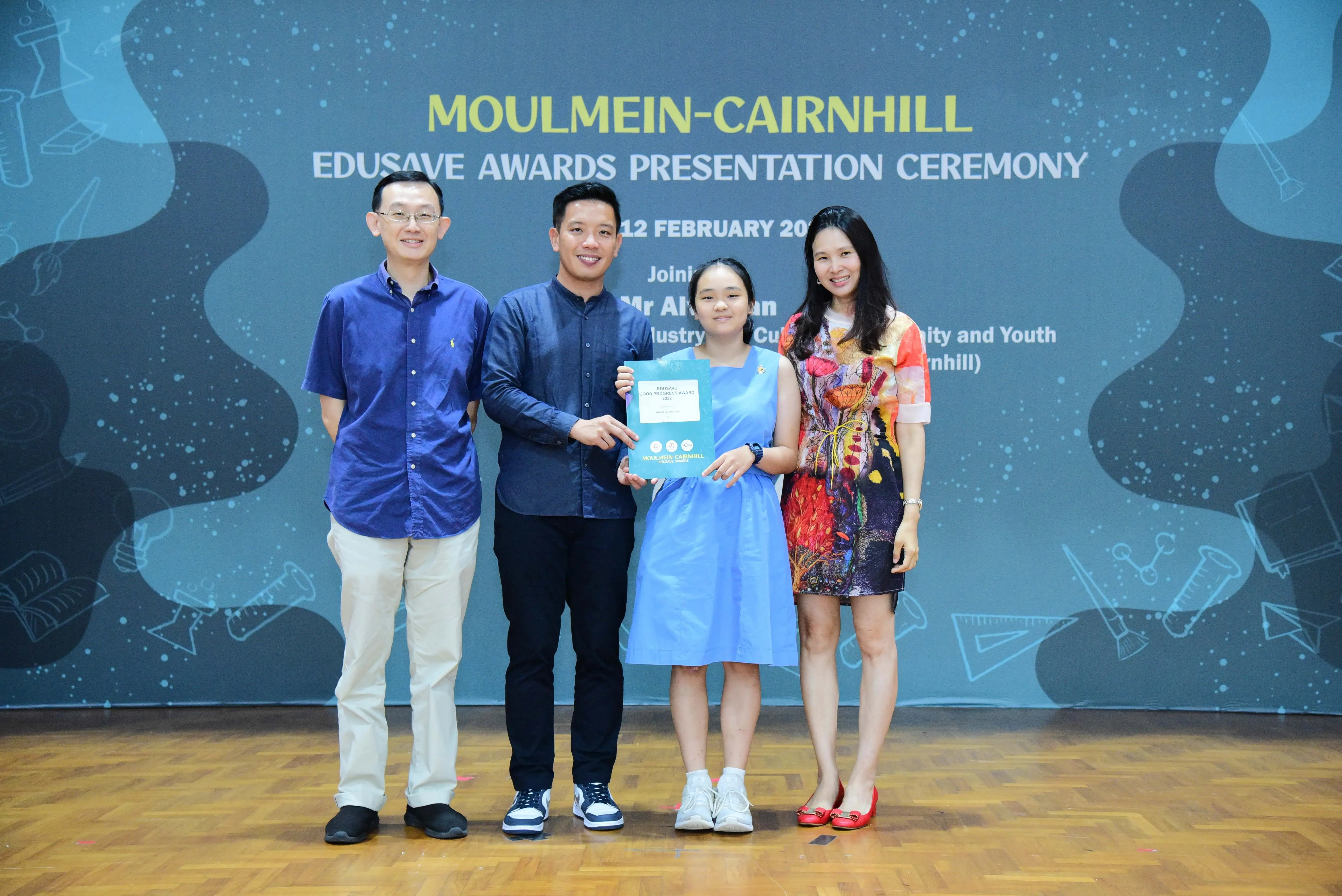 Four people pose on a stage at the Moulmein-Cairnhill Edusave Awards Presentation Ceremony, with a girl holding a certificate.