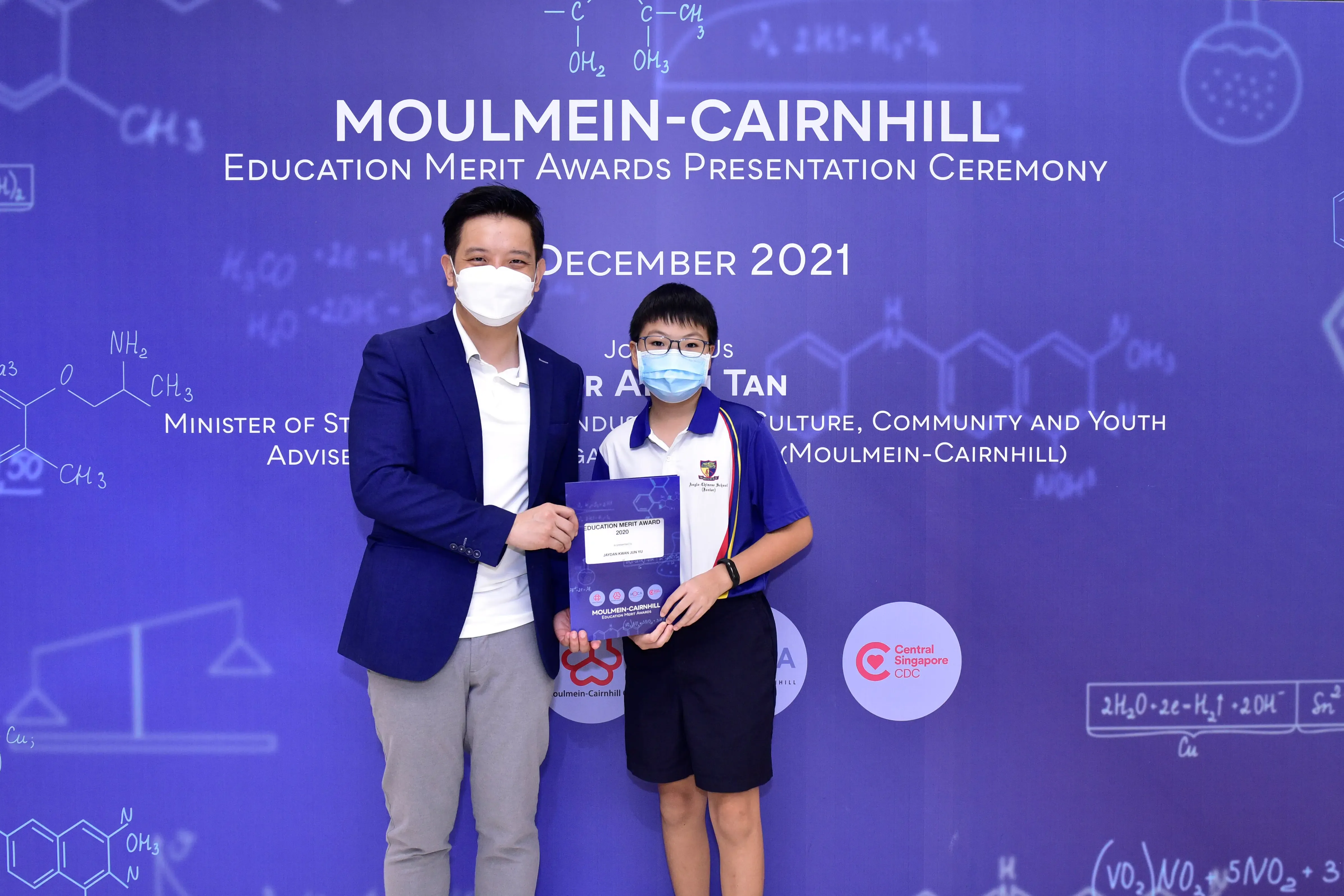 An adult and a young student wearing masks hold an award certificate at the Moulmein-Cairnhill Education Merit Awards December 2021.