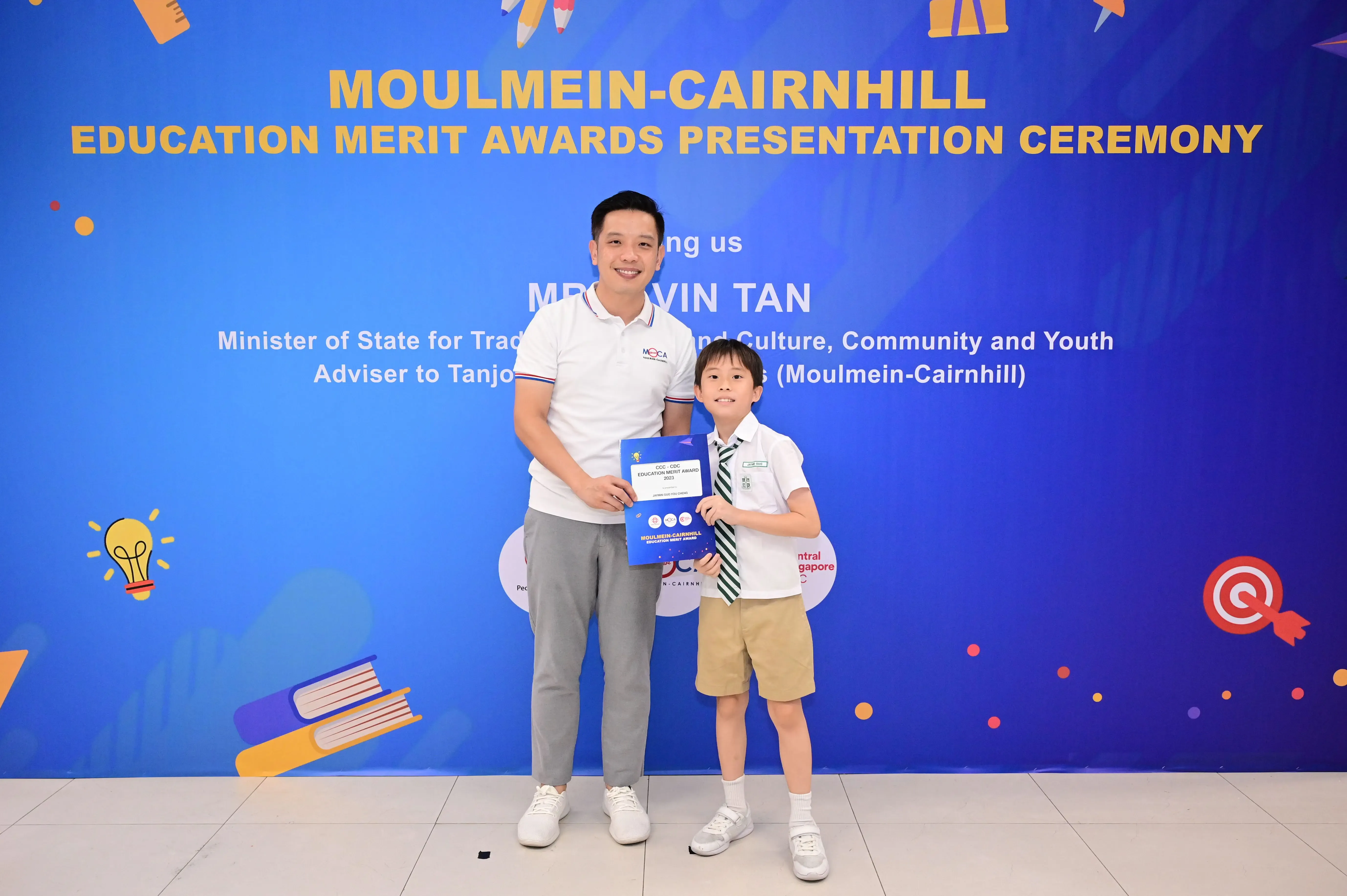 A man and a young boy hold a blue award certificate at the Moulmein-Cairnhill Education Merit Awards Ceremony.