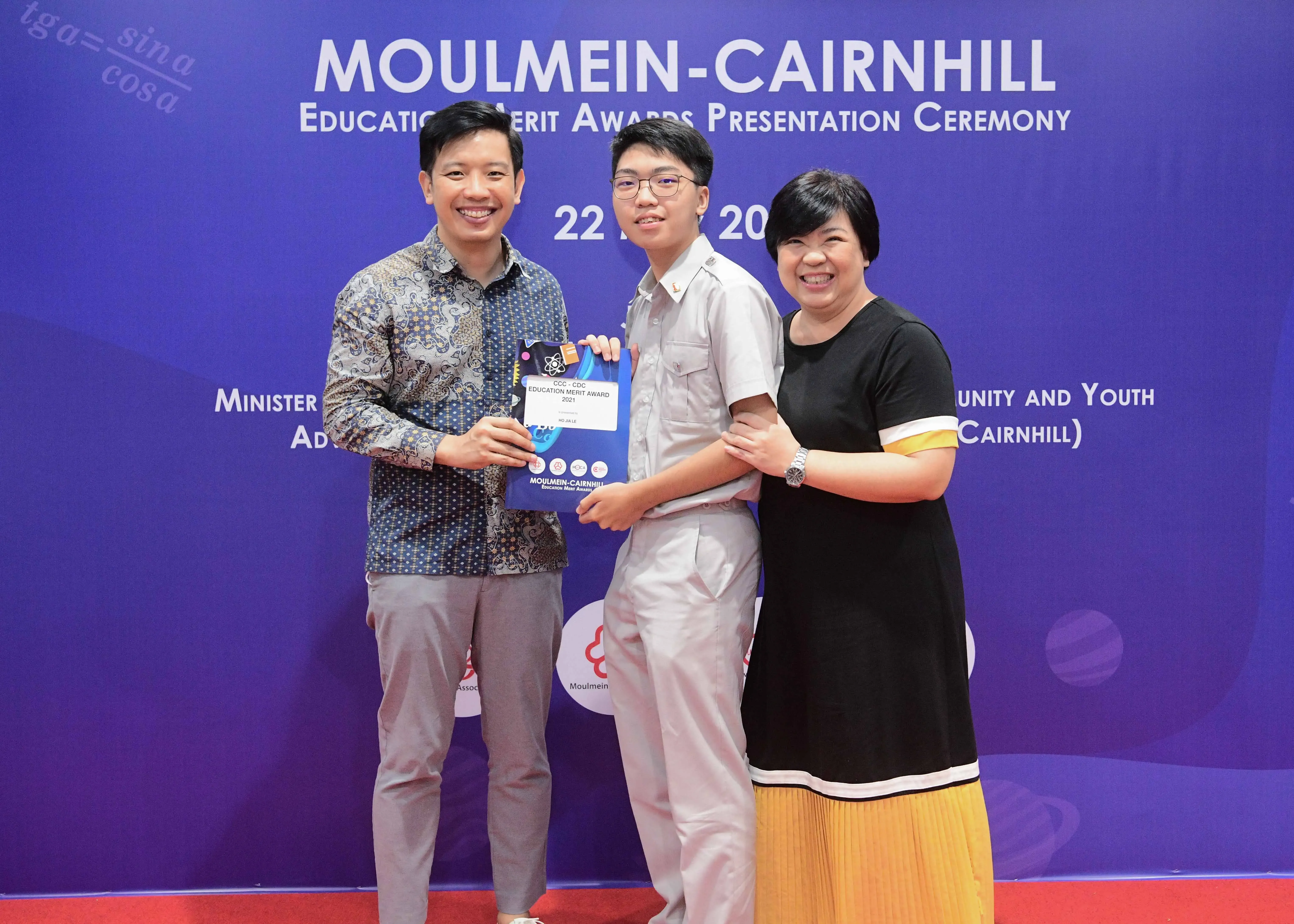 Three smiling people pose together at the Moulmein-Cairnhill Education Merit Awards Presentation Ceremony, holding a certificate.