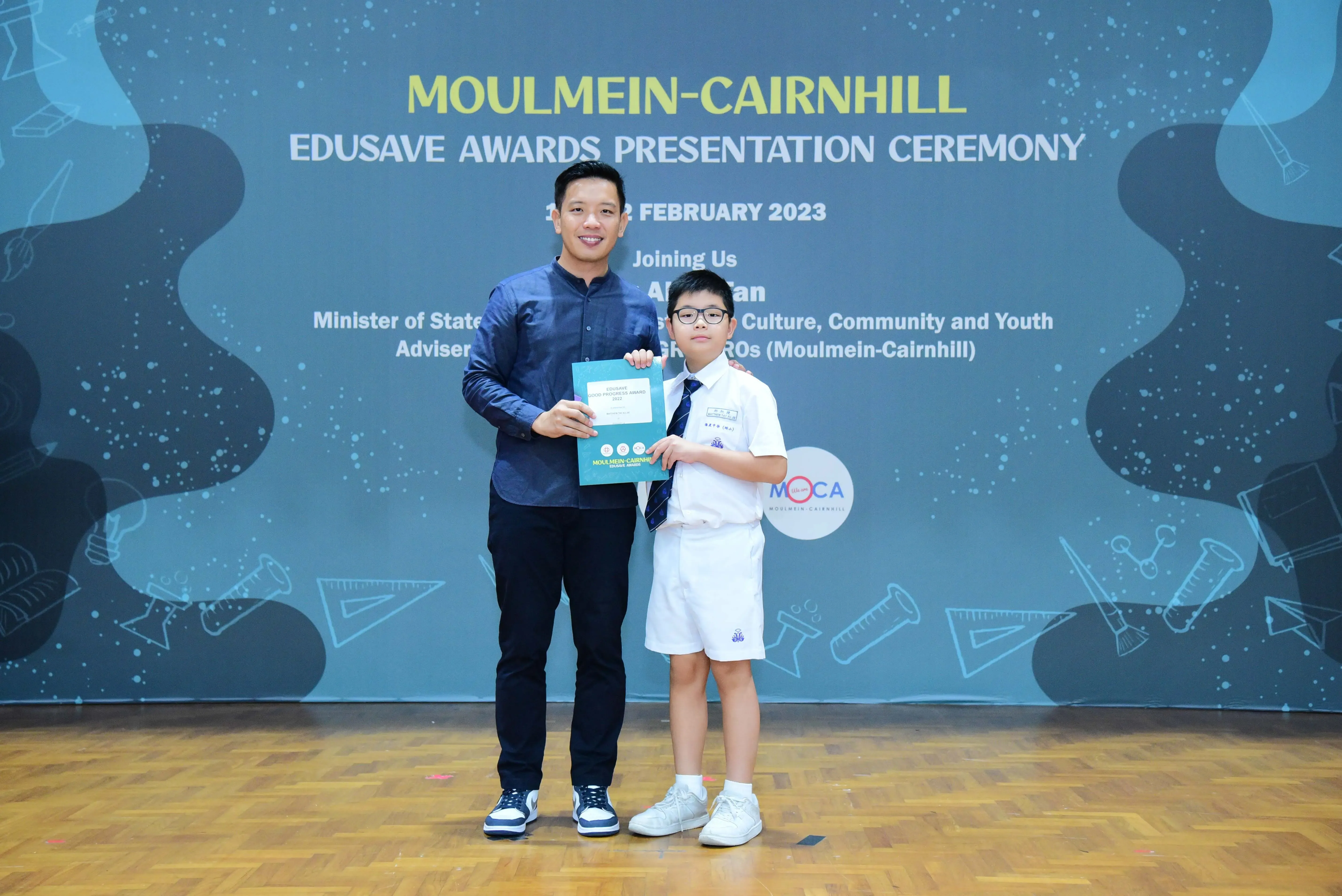 An adult and a young student in uniform hold a certificate at the Moulmein-Cairnhill Edusave Awards Ceremony, February 2023.