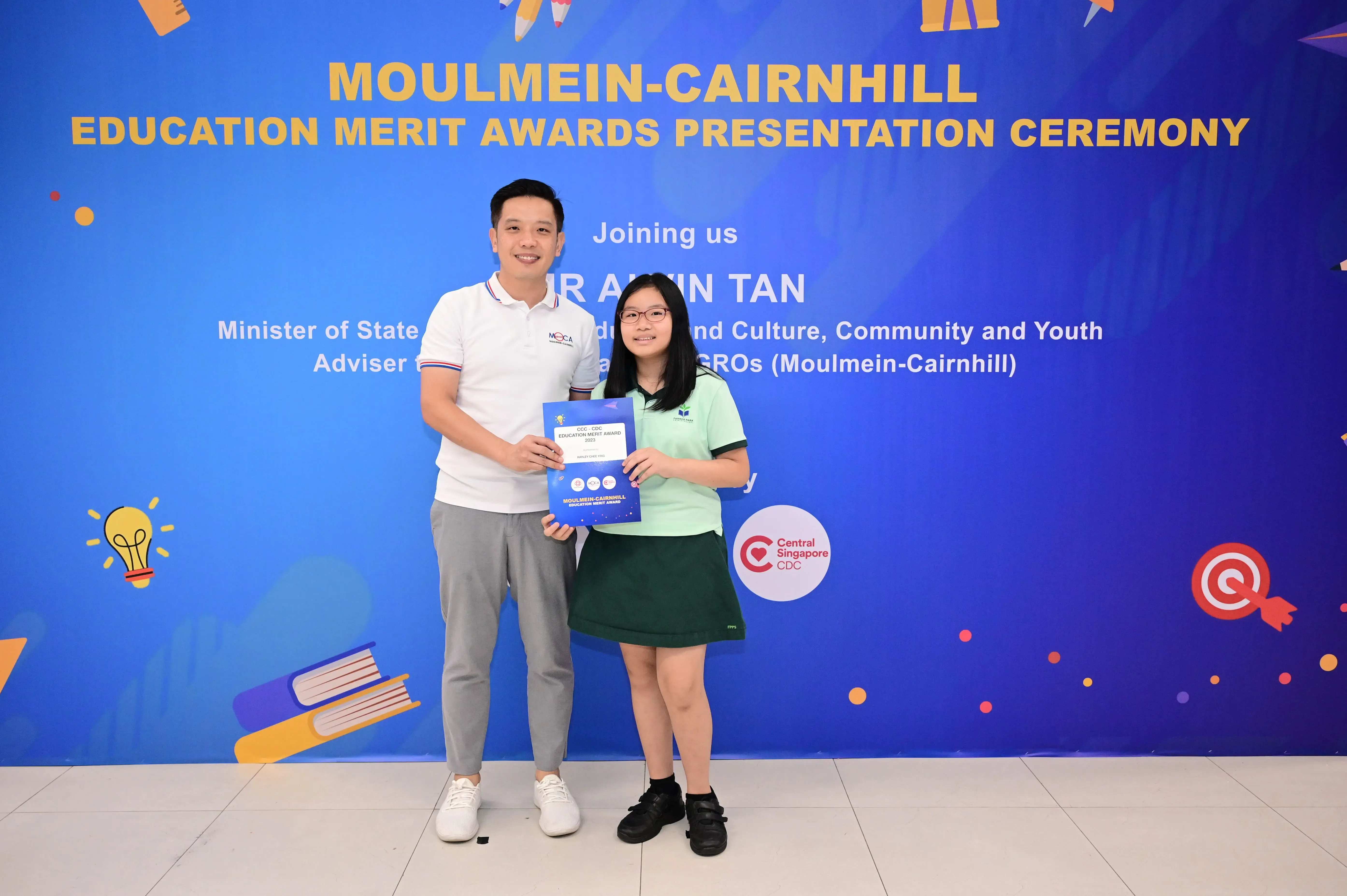 A man and a young girl hold an award certificate in front of a Moulmein-Cairnhill Education Merit Awards banner.