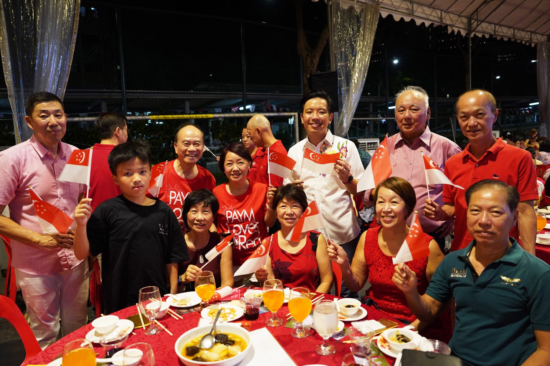 A group of smiling people in red outfits wave Singapore flags at a festive outdoor dinner table at night.