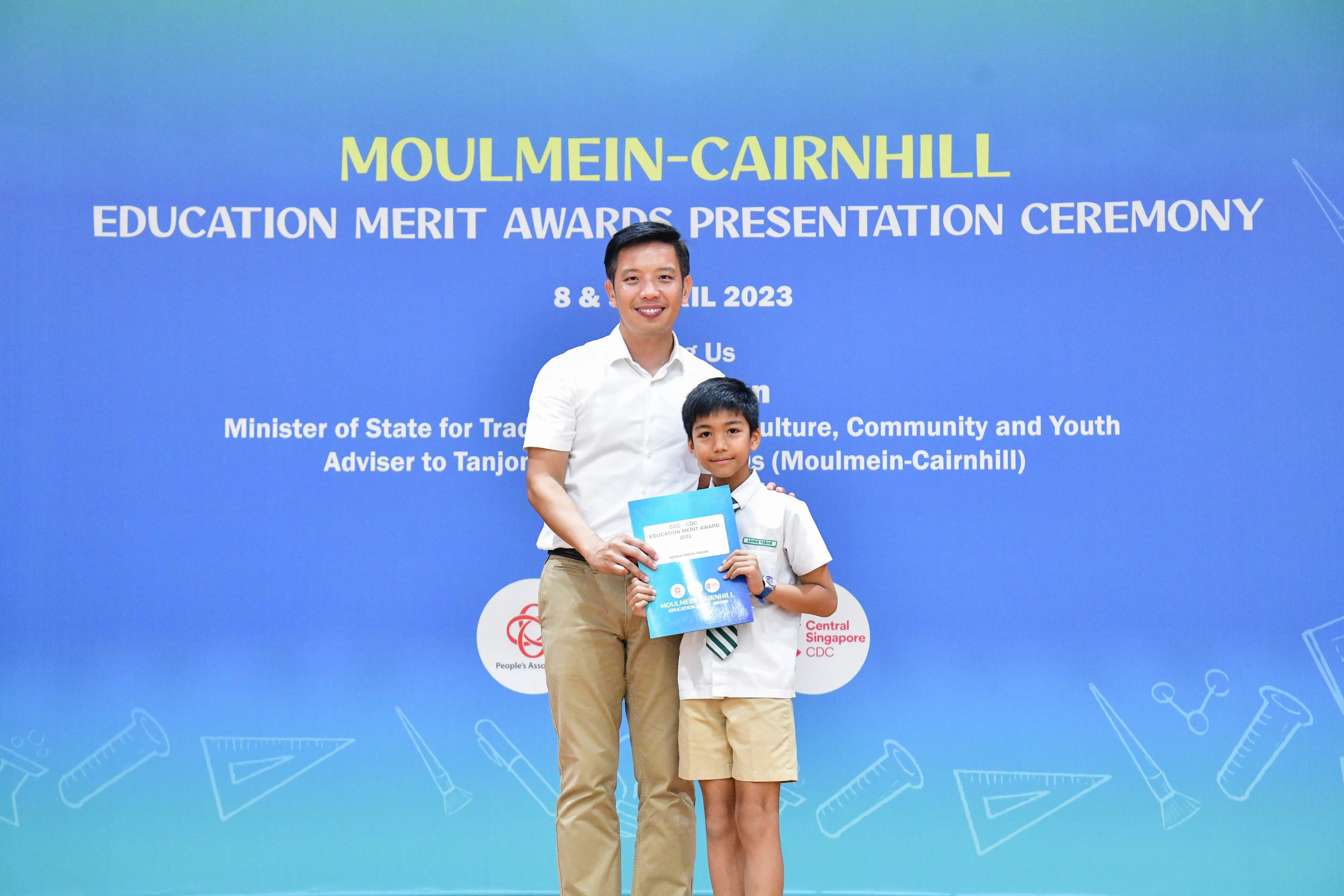 Adult man and young boy pose together holding an award booklet at the Moulmein-Cairnhill Education Merit Awards 2023.
