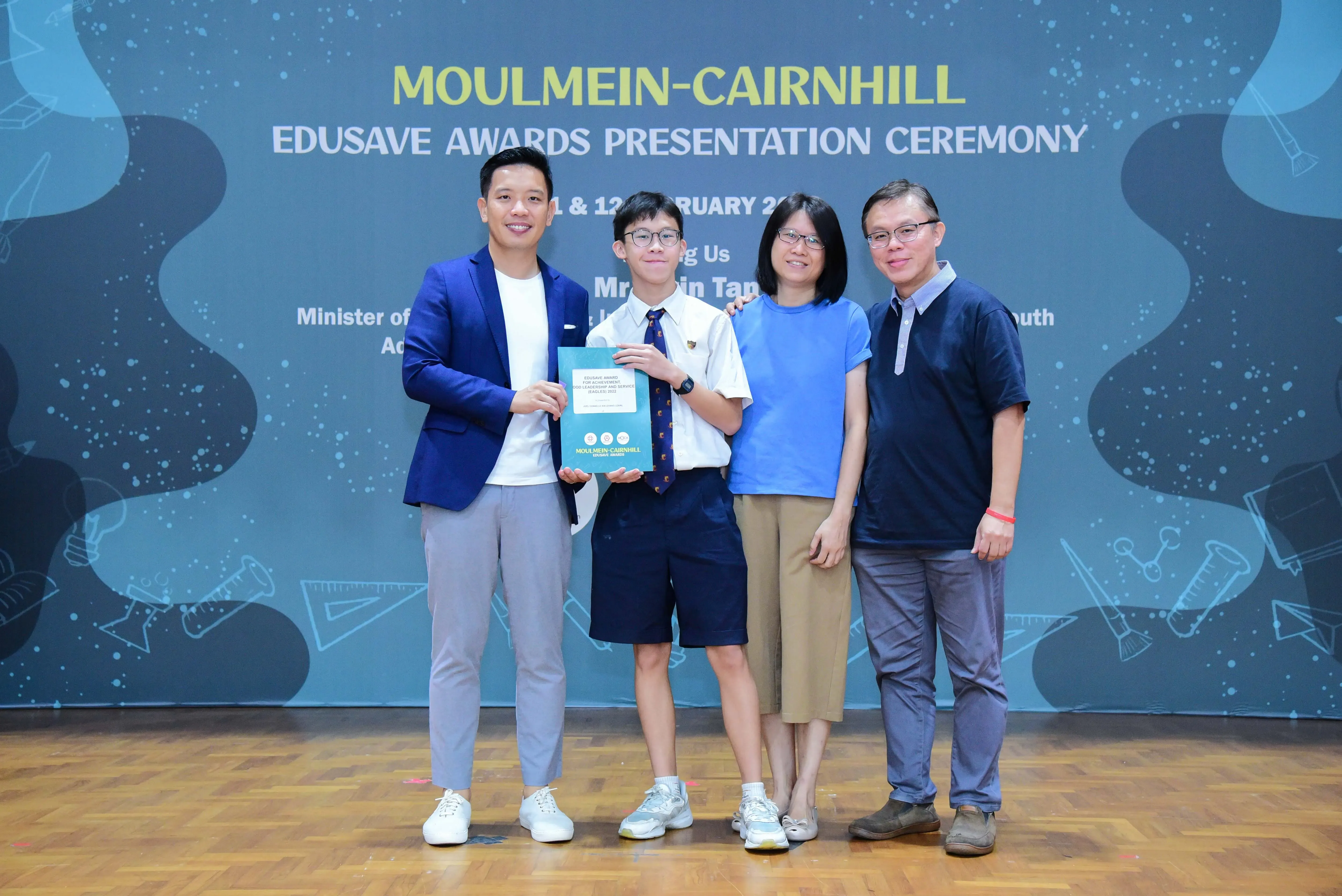 Four people pose on a stage at the Moulmein-Cairnhill Edusave Awards Presentation Ceremony, holding a certificate.