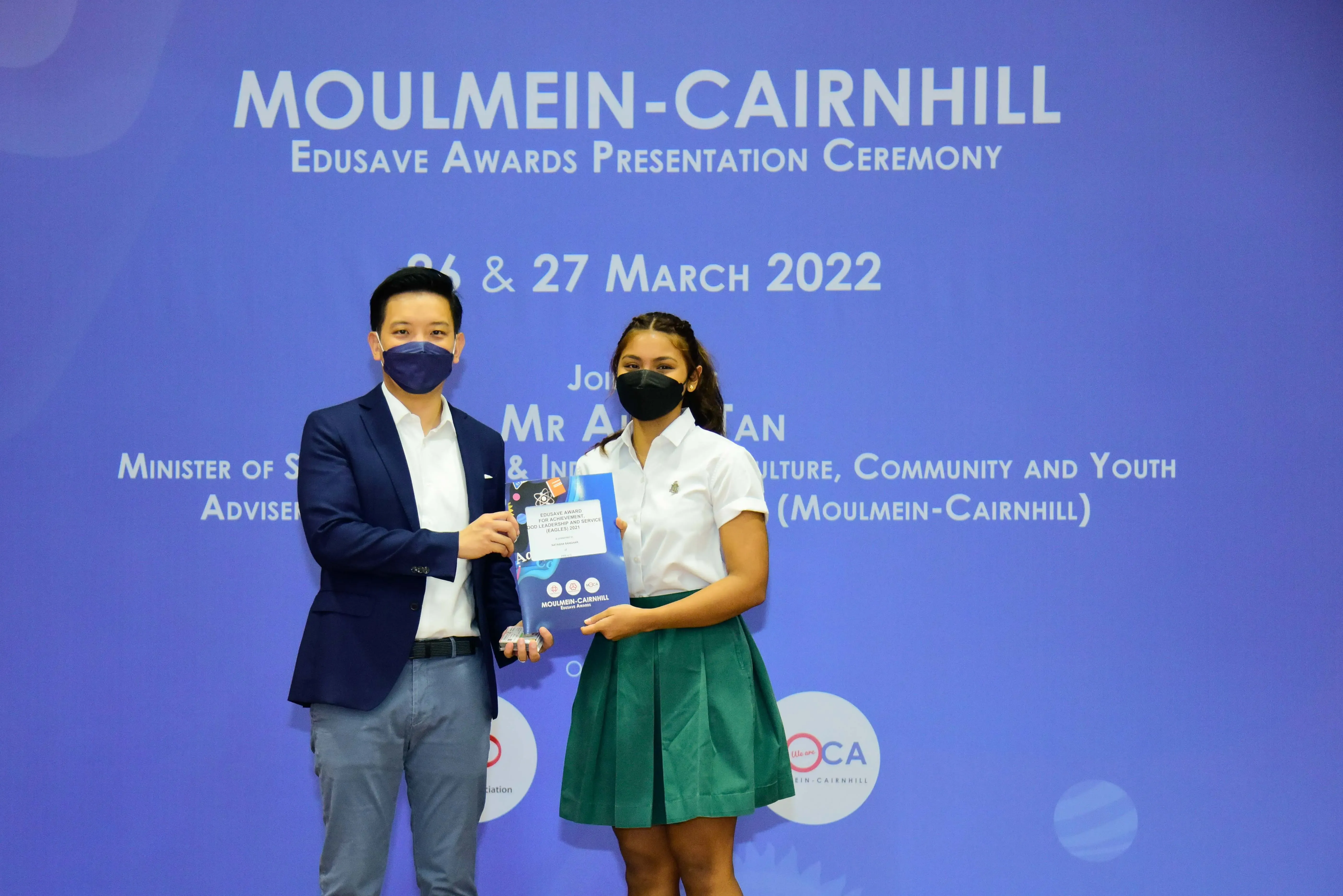 Two masked people hold an award at the Moulmein-Cairnhill Edusave Awards Presentation Ceremony, March 2022.
