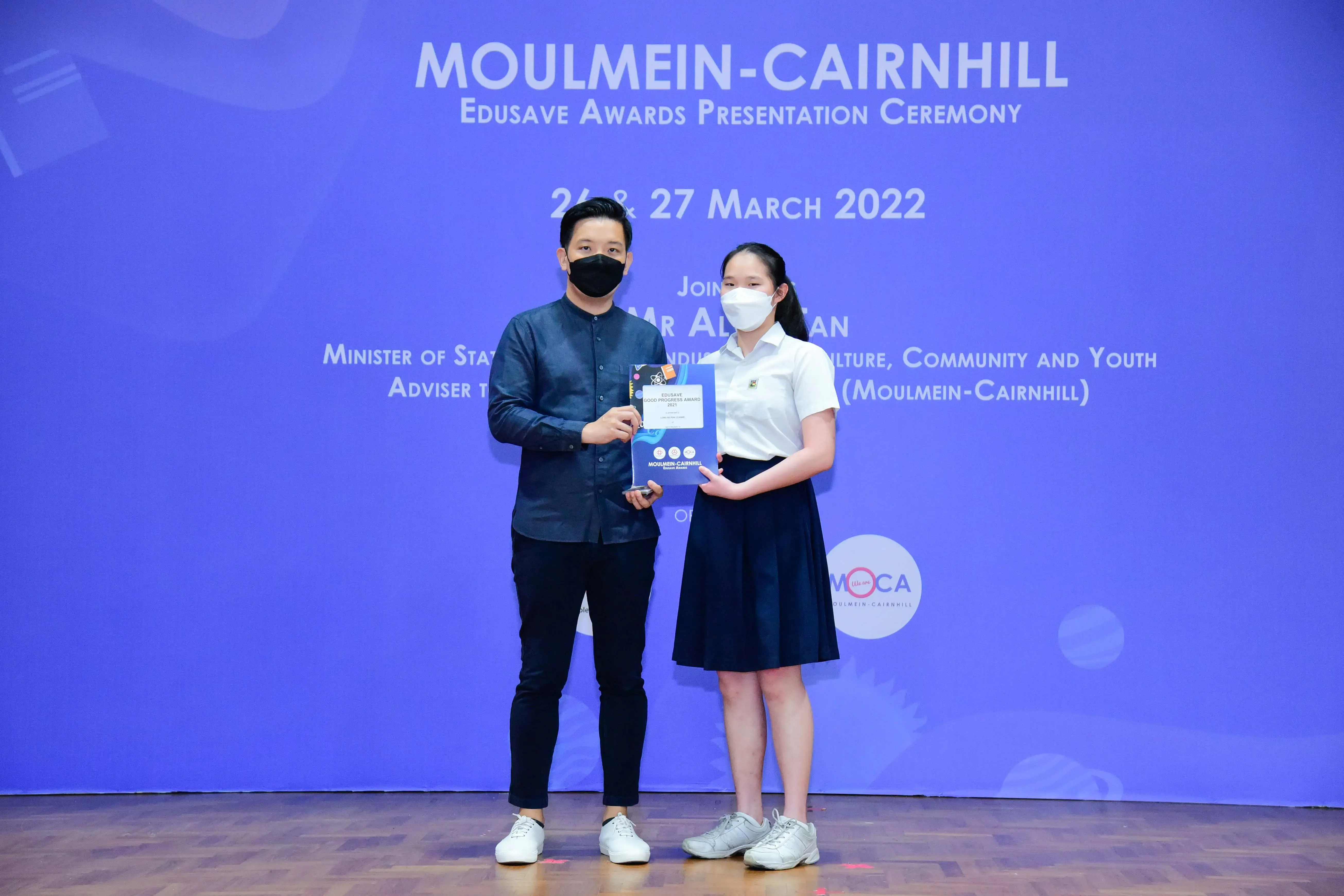Two masked individuals hold a certificate at the Moulmein-Cairnhill Edusave Awards Presentation Ceremony, March 2022.