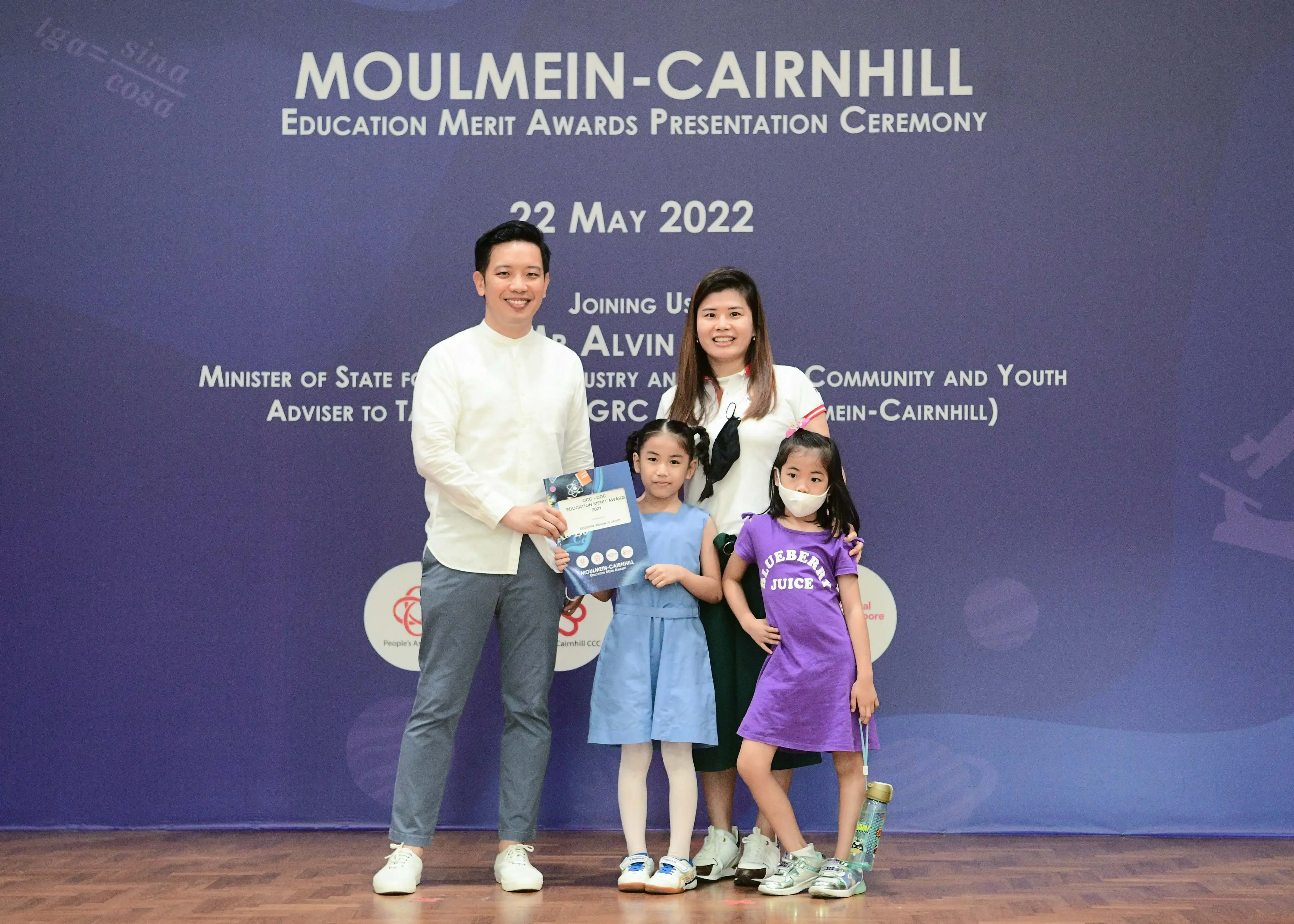 A family of four poses in front of the Moulmein-Cairnhill Education Merit Awards banner dated 22 May 2022.