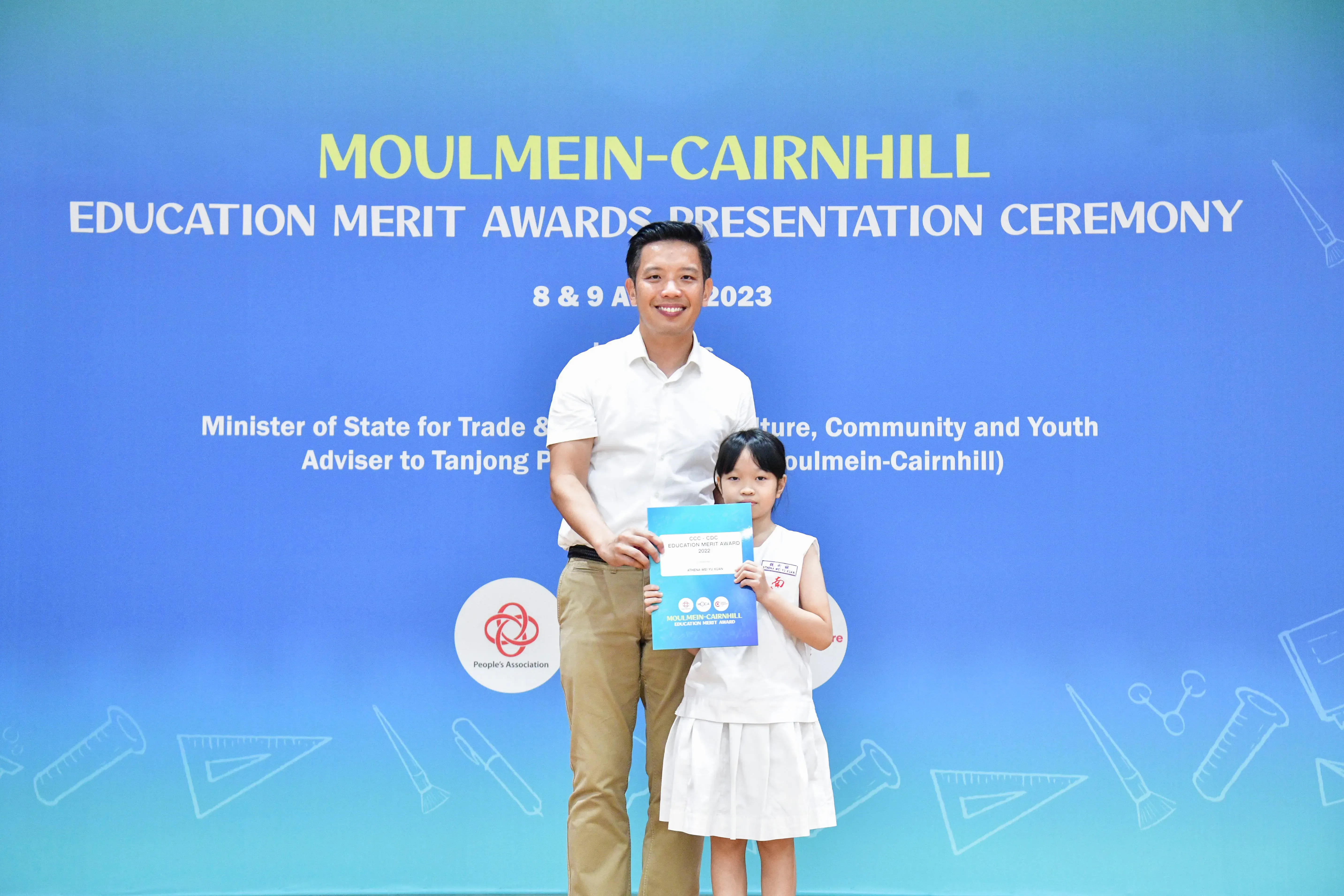 A man and a young girl holding an award certificate stand before a Moulmein-Cairnhill Education Merit Awards backdrop.