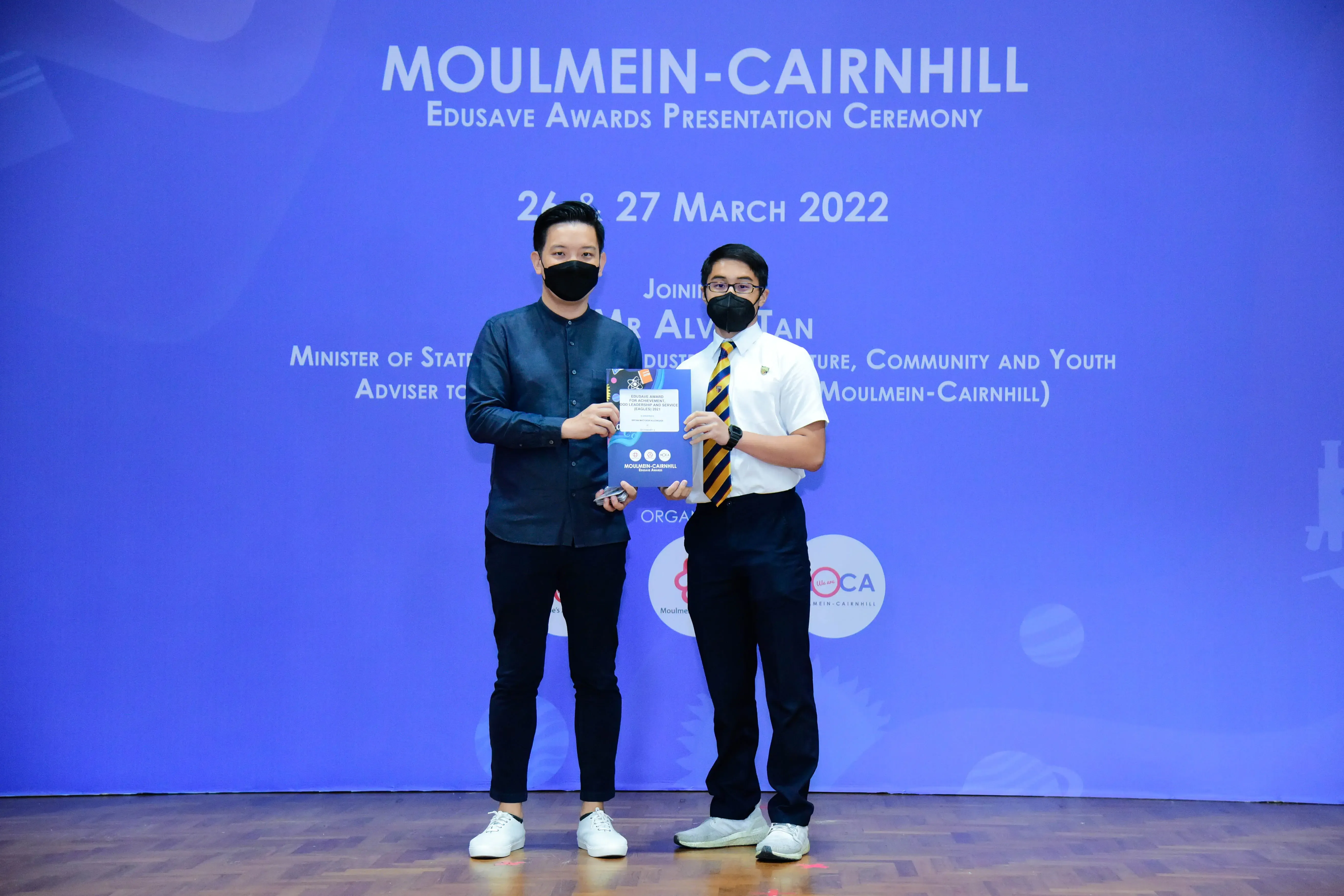 Two masked men hold an award at the Moulmein-Cairnhill Edusave Awards Presentation Ceremony, March 2022.